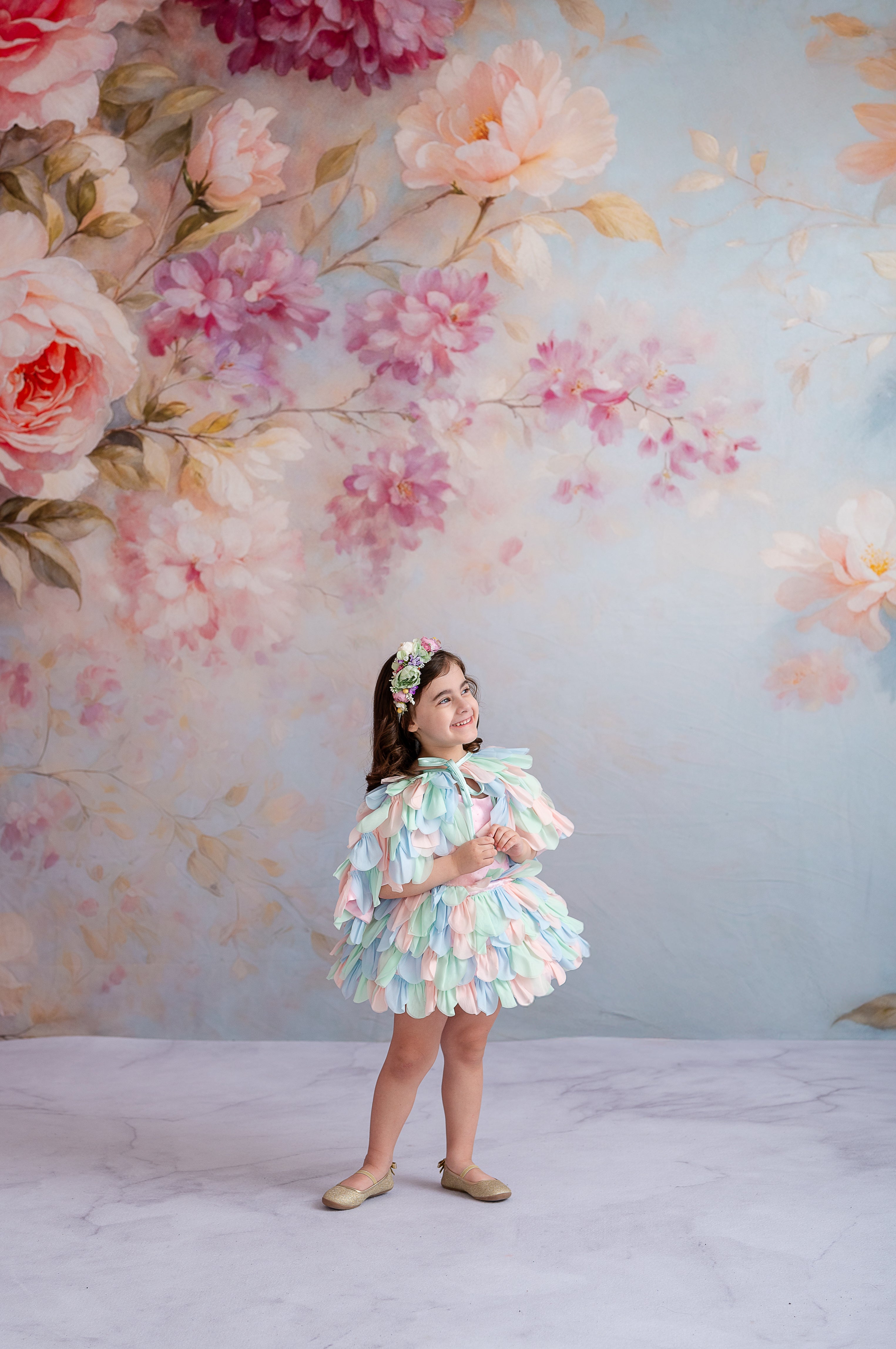 "Elegant young girl in a whimsical, petal-inspired dress stands before a painted floral backdrop."