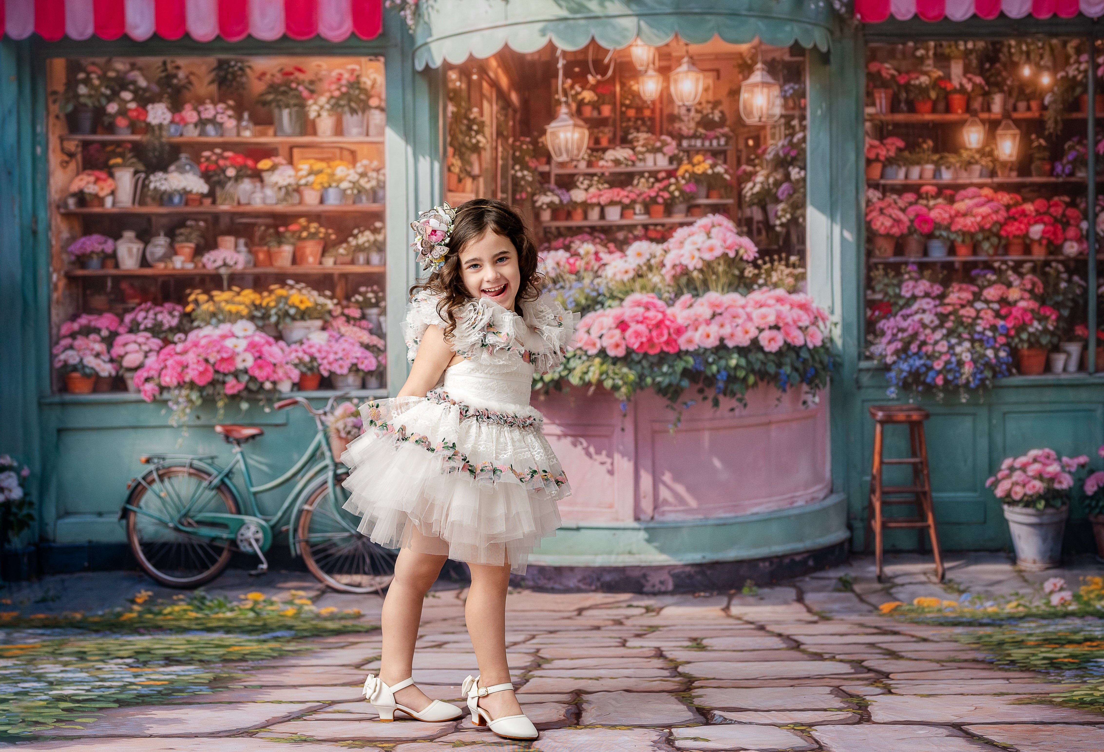 "A young girl twirls in a layered white dress with floral accents, set against a backdrop of blooming flowers and pastel storefronts."