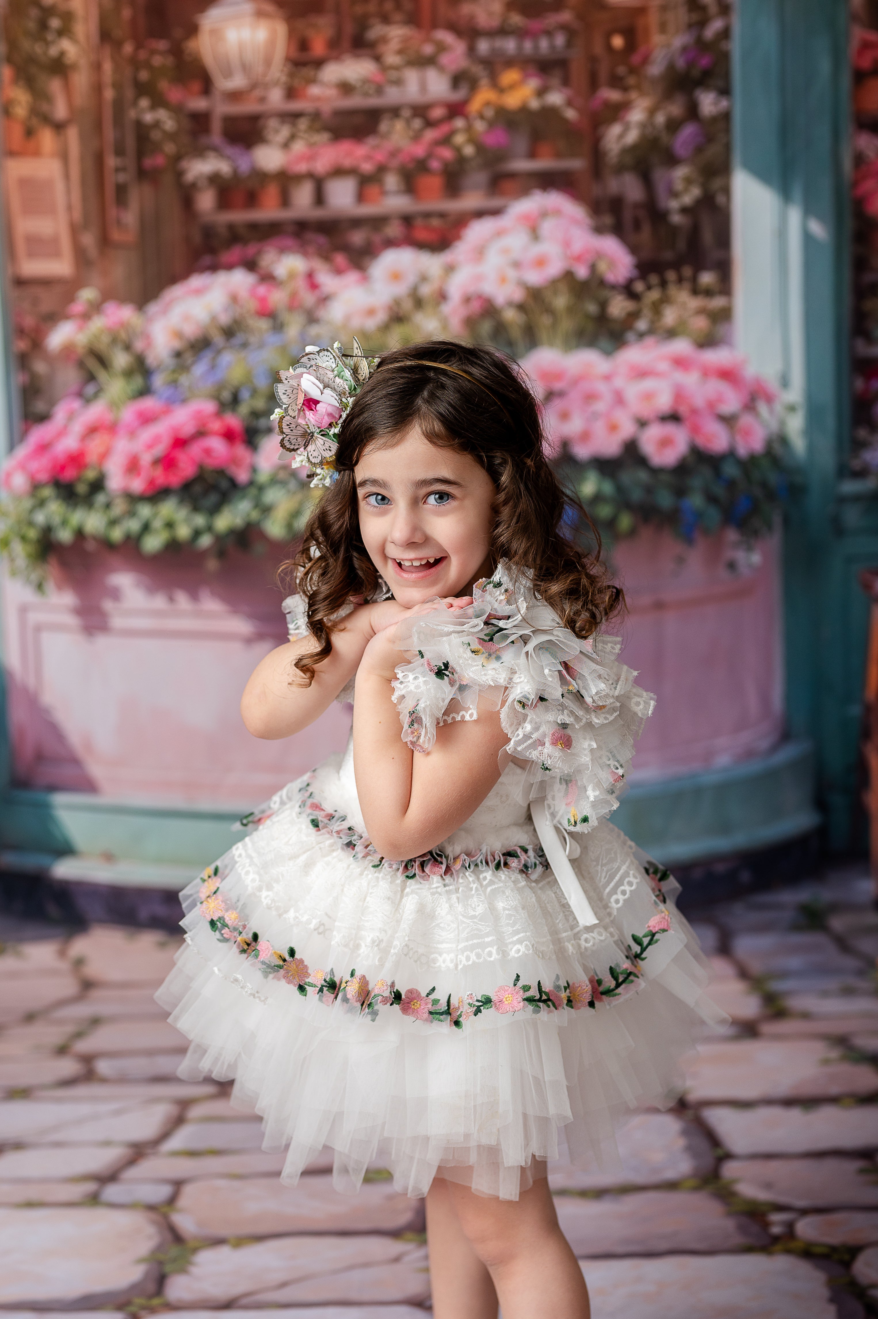 "A cheerful child in a white and floral tulle gown stands on a cobblestone street with a charming flower boutique in the background."