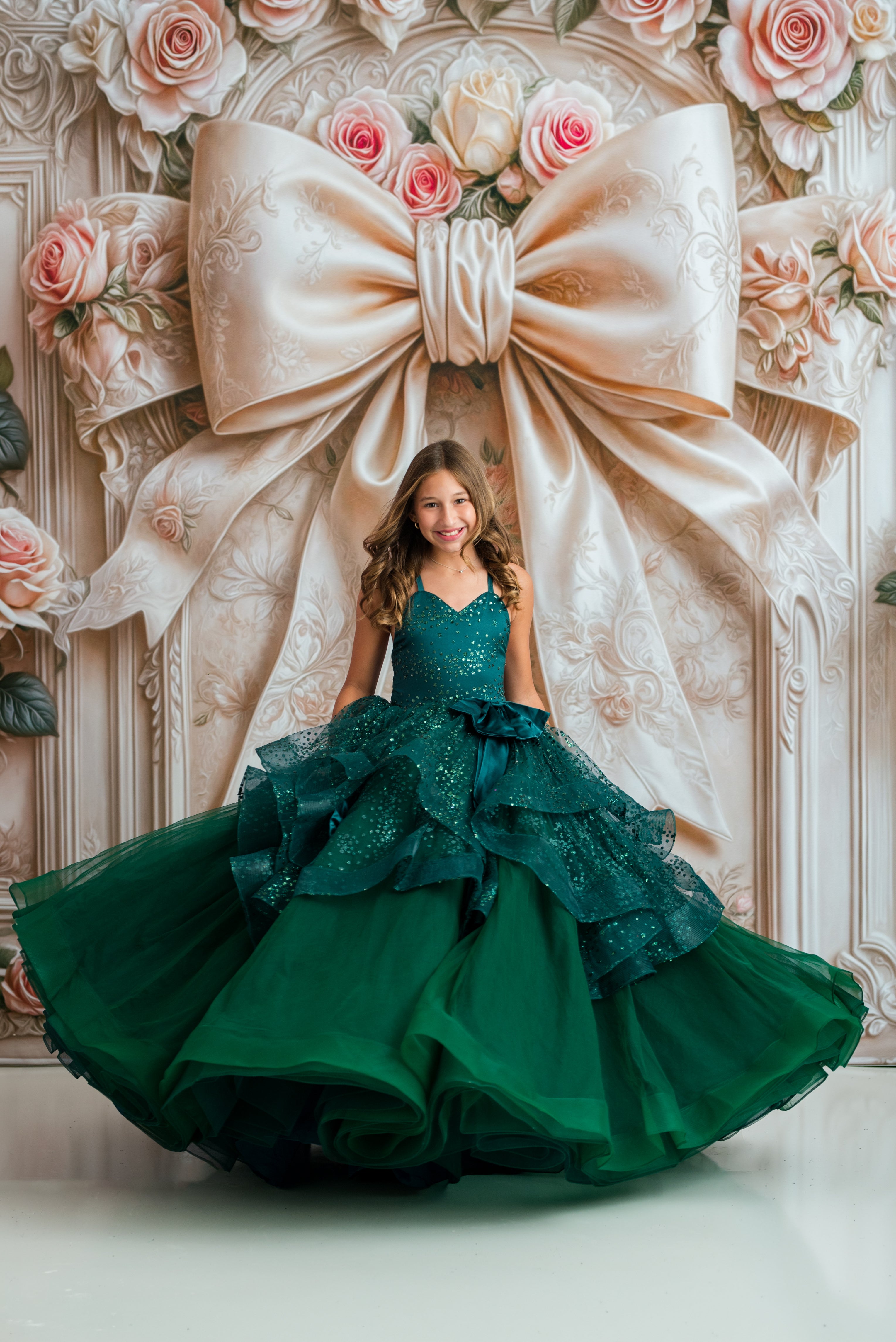 A young girl wearing a voluminous emerald green ball gown with a sparkling bodice and layered ruffles, posing in front of a decorative backdrop featuring a giant cream satin bow and pink roses.
