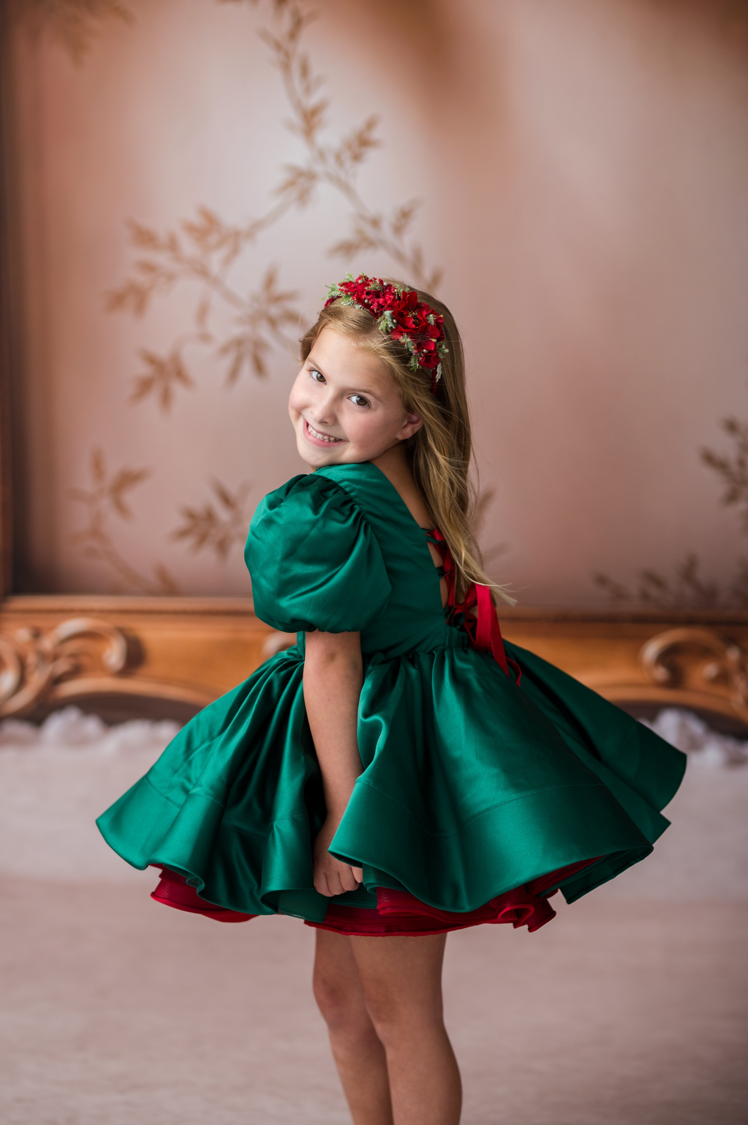 A young girl with blonde hair wearing a red floral headband, looking back over her shoulder while wearing a shiny emerald green dress with a red underlayer and red sash, standing in front of a warm beige wall with leaf patterns.