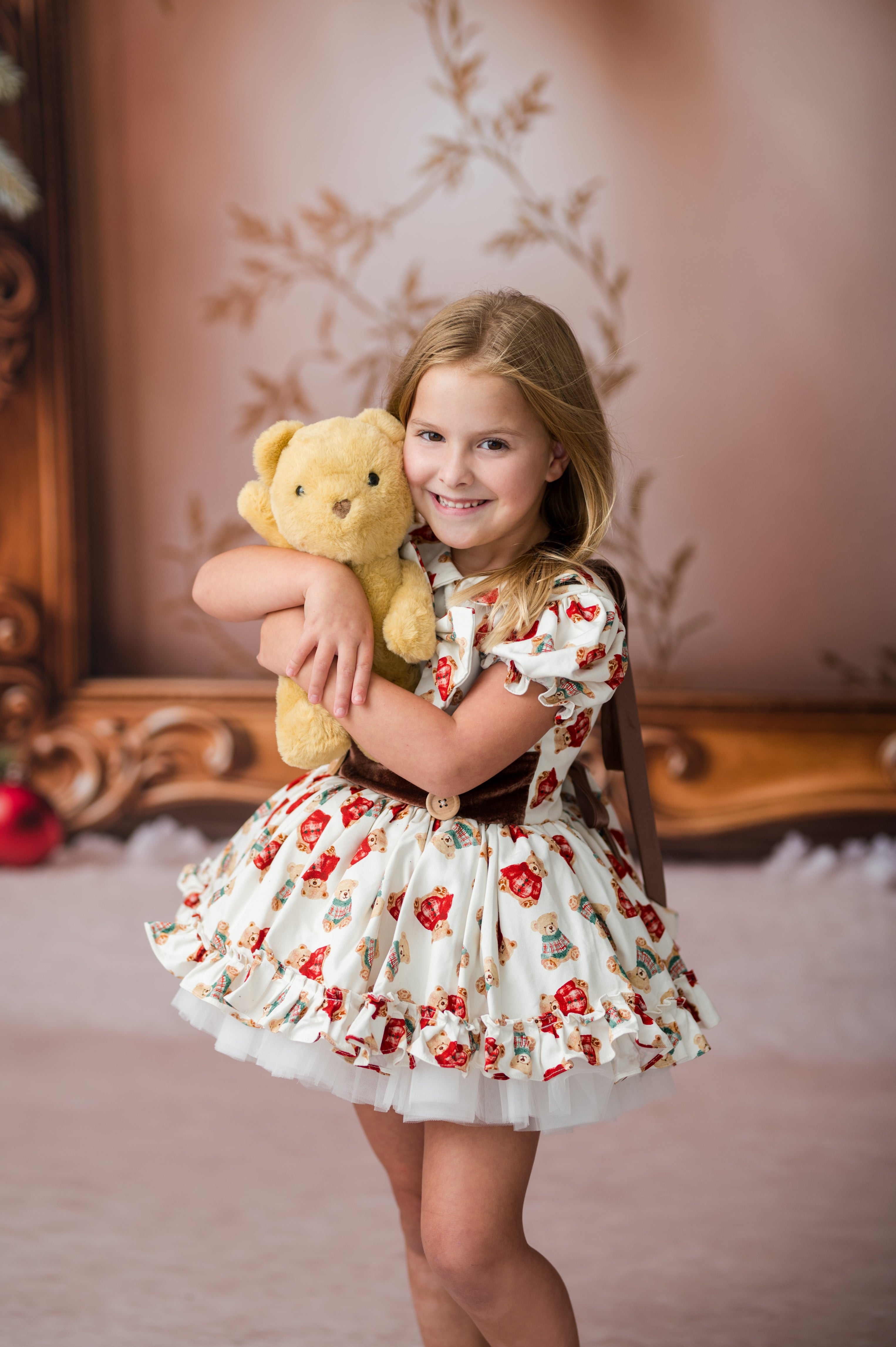A young girl with blonde hair smiling while holding a yellow teddy bear, wearing a white ruffled dress with a red and green floral pattern and a dark brown sash, posed in front of a vintage-style mirror and pink wall.