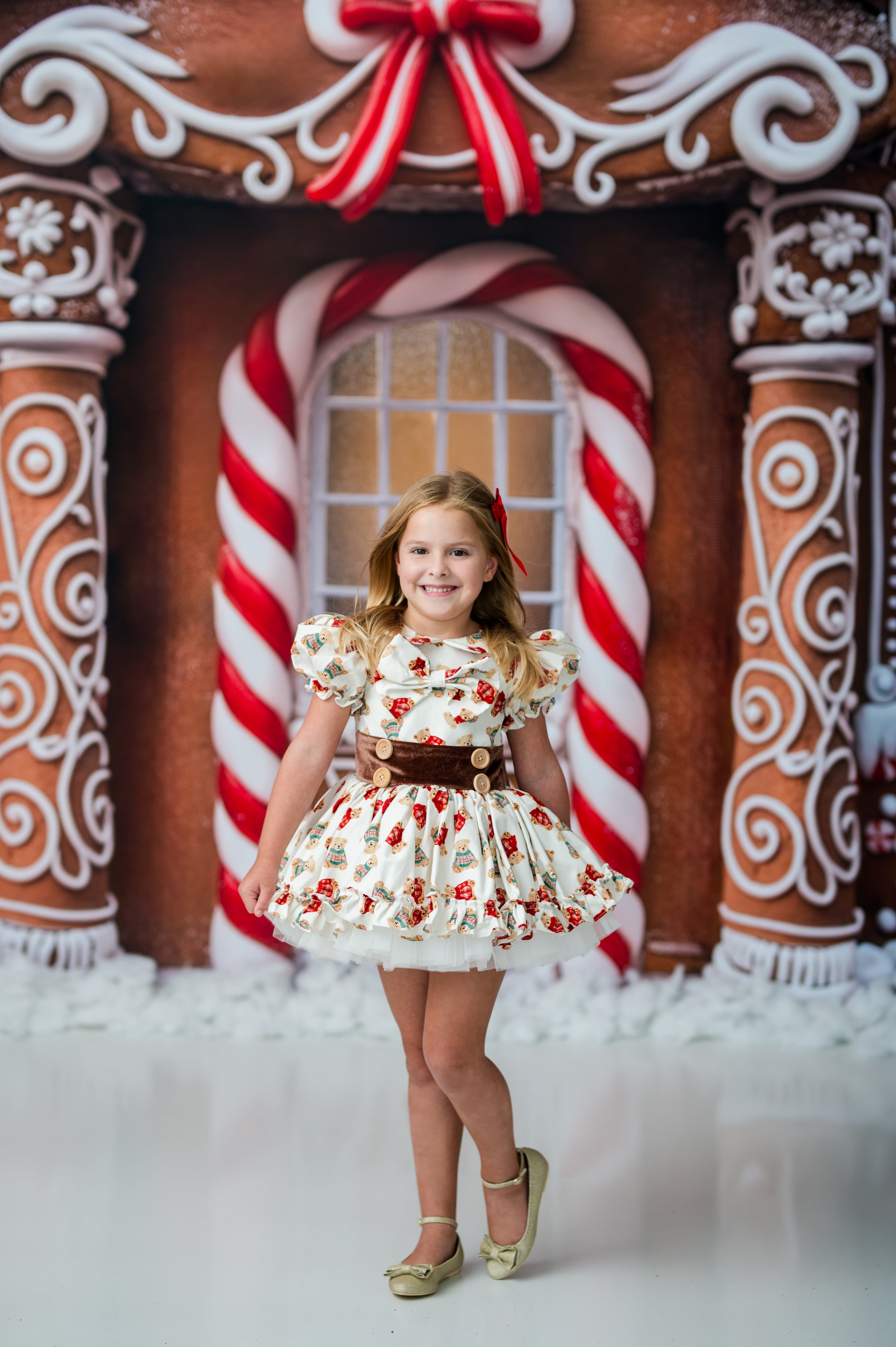 A young girl stands in front of a gingerbread house backdrop wearing a short white dress with red floral print, a brown waistband, and gold shoes.
