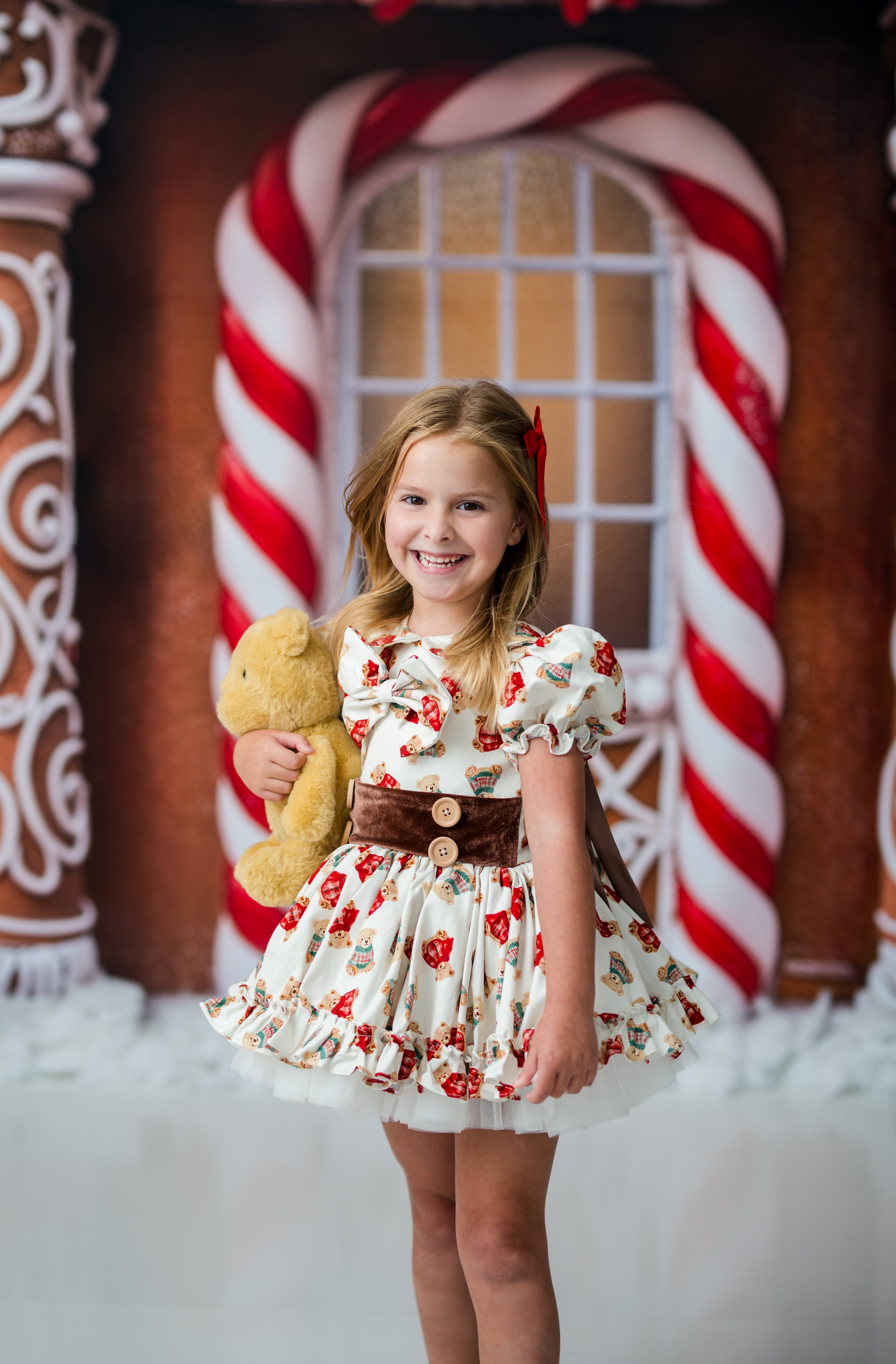 A young girl stands smiling in front of a gingerbread-themed backdrop with candy cane arches, wearing a white dress with red floral print, a brown waist belt, and holding a yellow teddy bear.