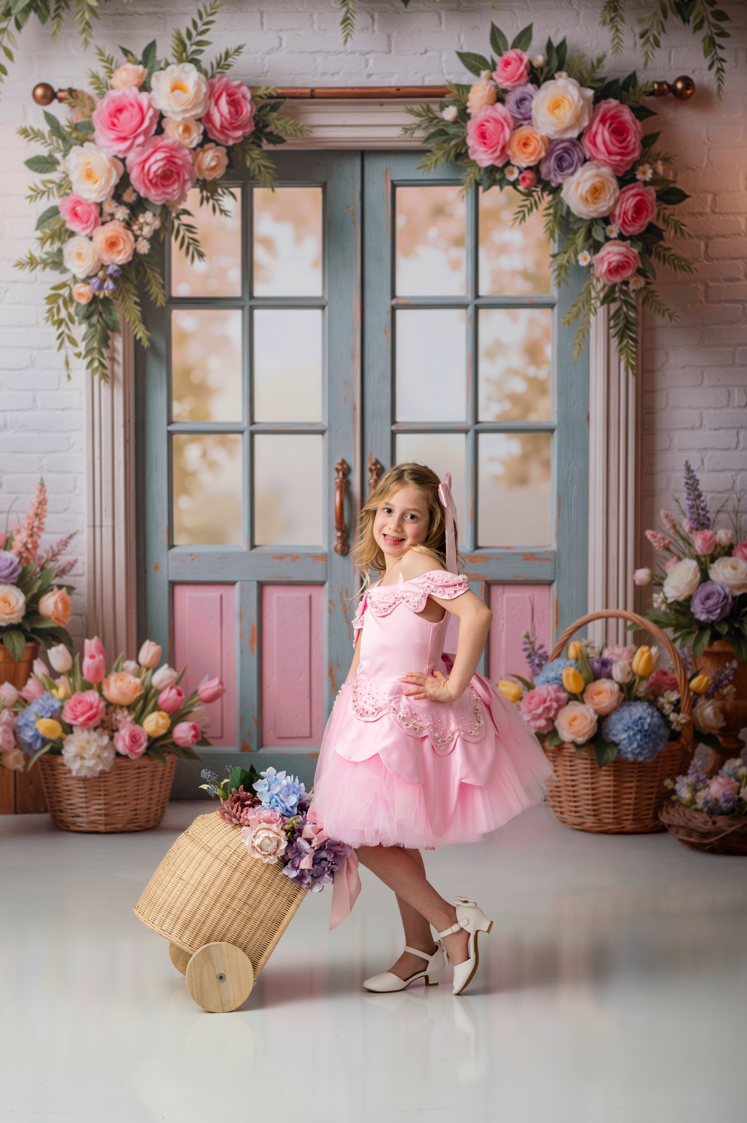 Young girl in a pink dress standing in front of a decorated backdrop with flowers and a suitcase.