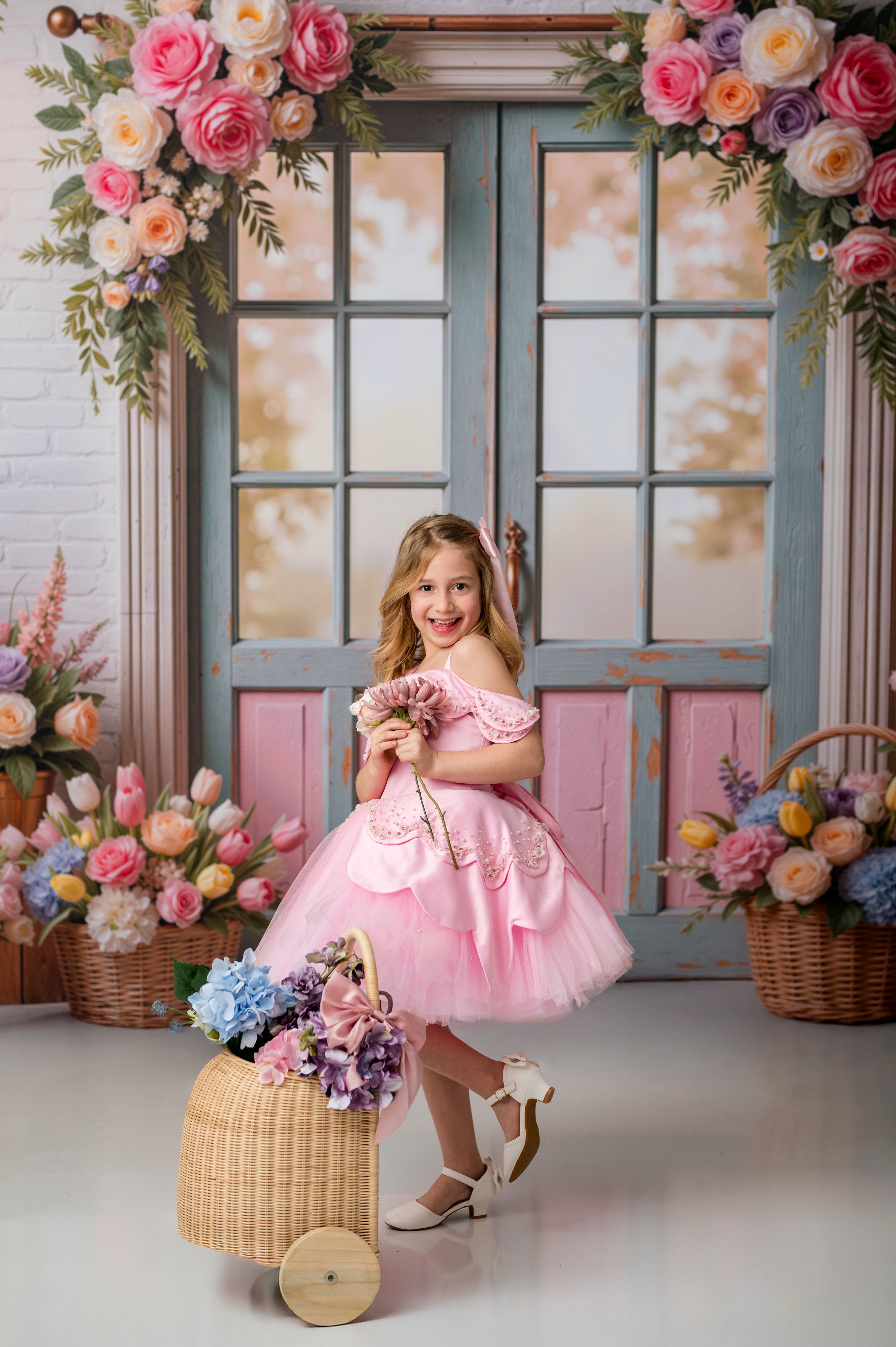 Young girl in a pink dress holding flowers, standing in front of a decorative floral arch and window.