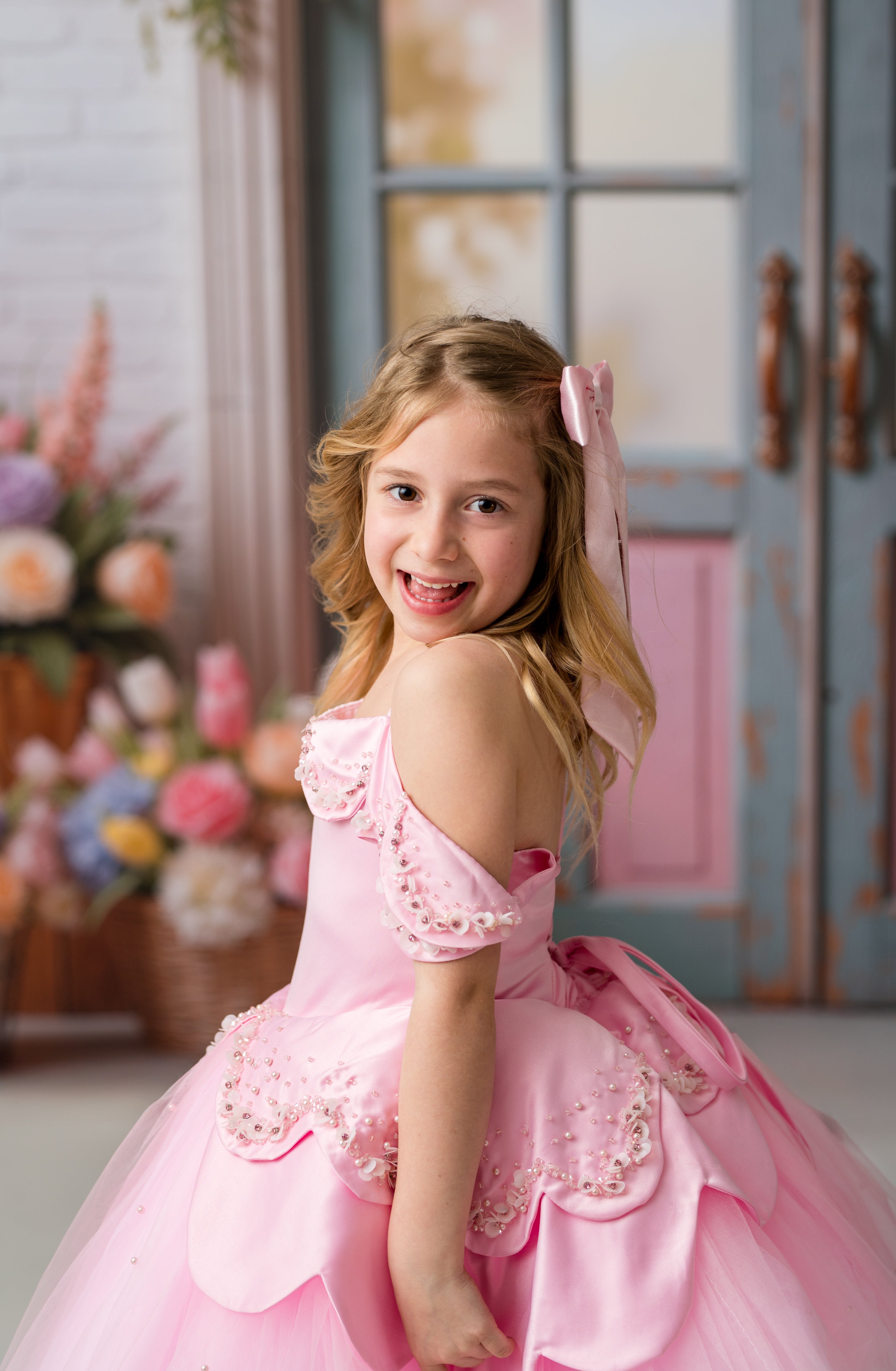 Young girl in a pink dress standing in a room with flowers and a door in the background