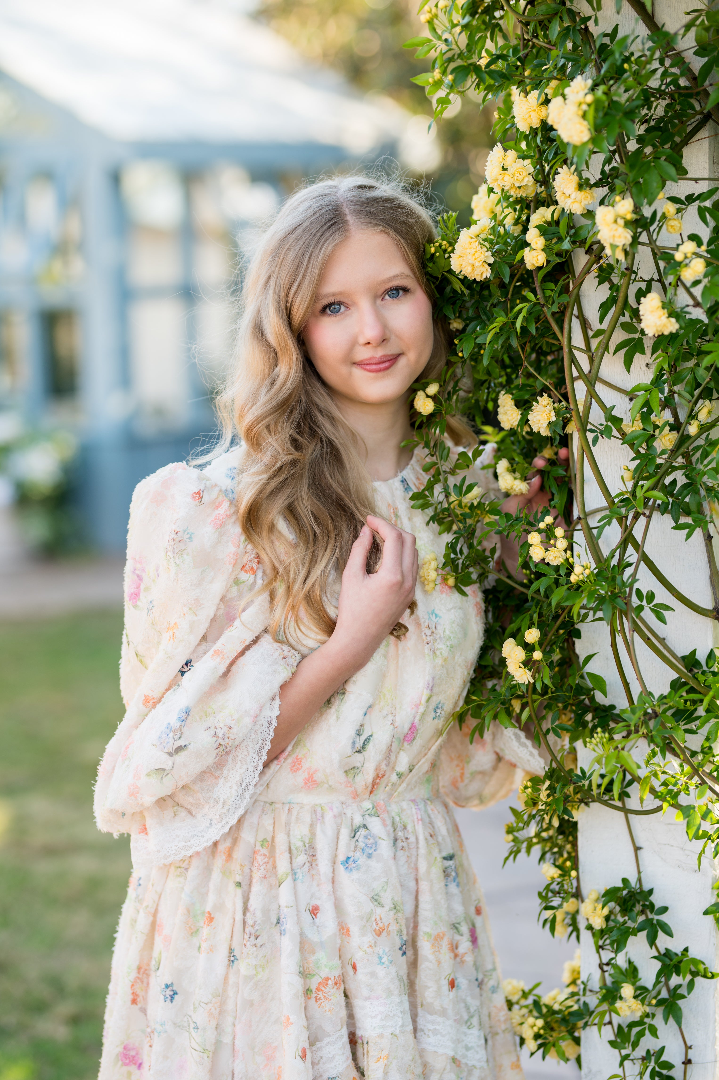 Young woman in a floral dress standing next to a wall with yellow flowers outdoors.