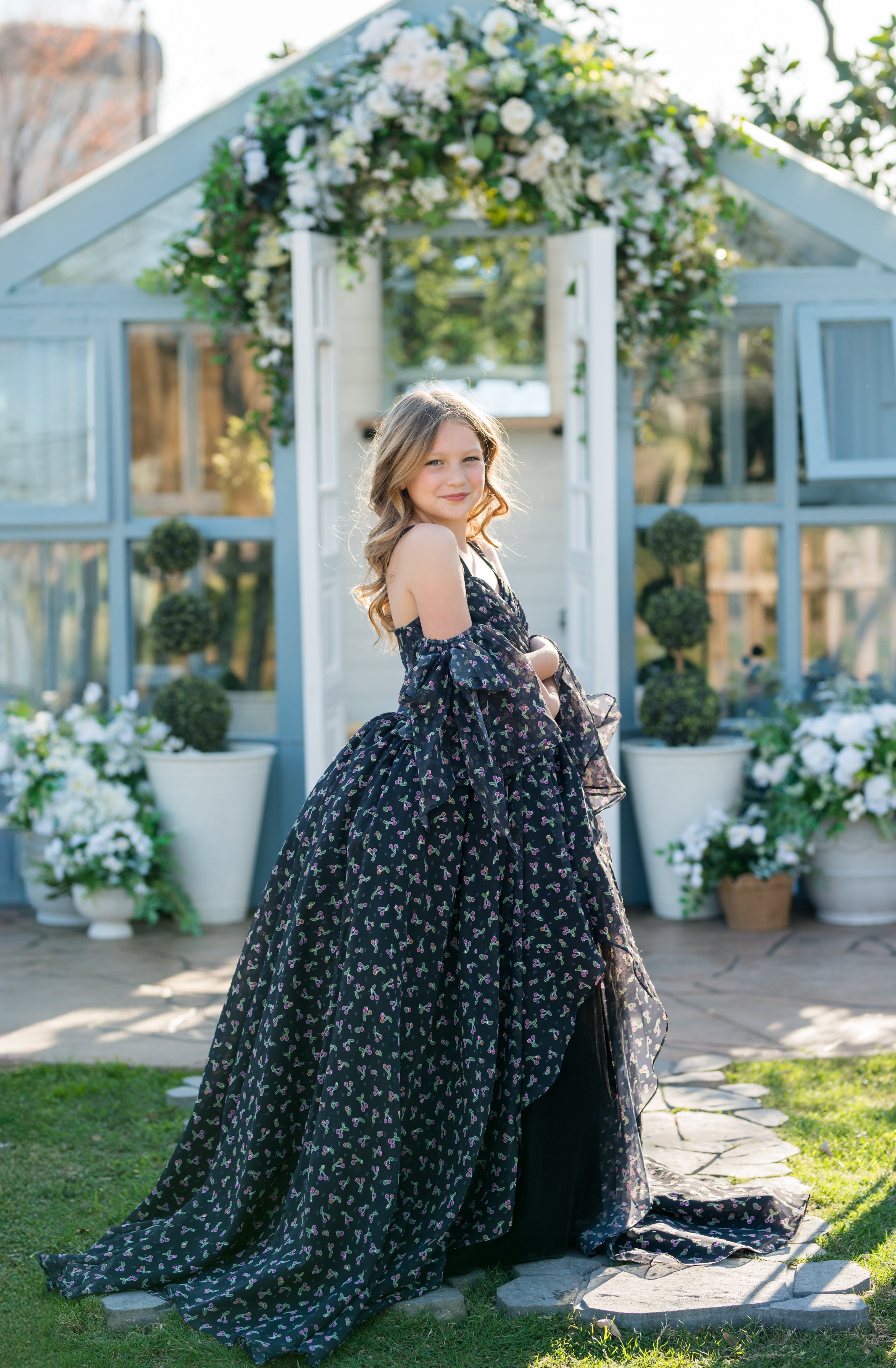 Young girl in a black floral dress standing in front of a decorative archway with flowers.