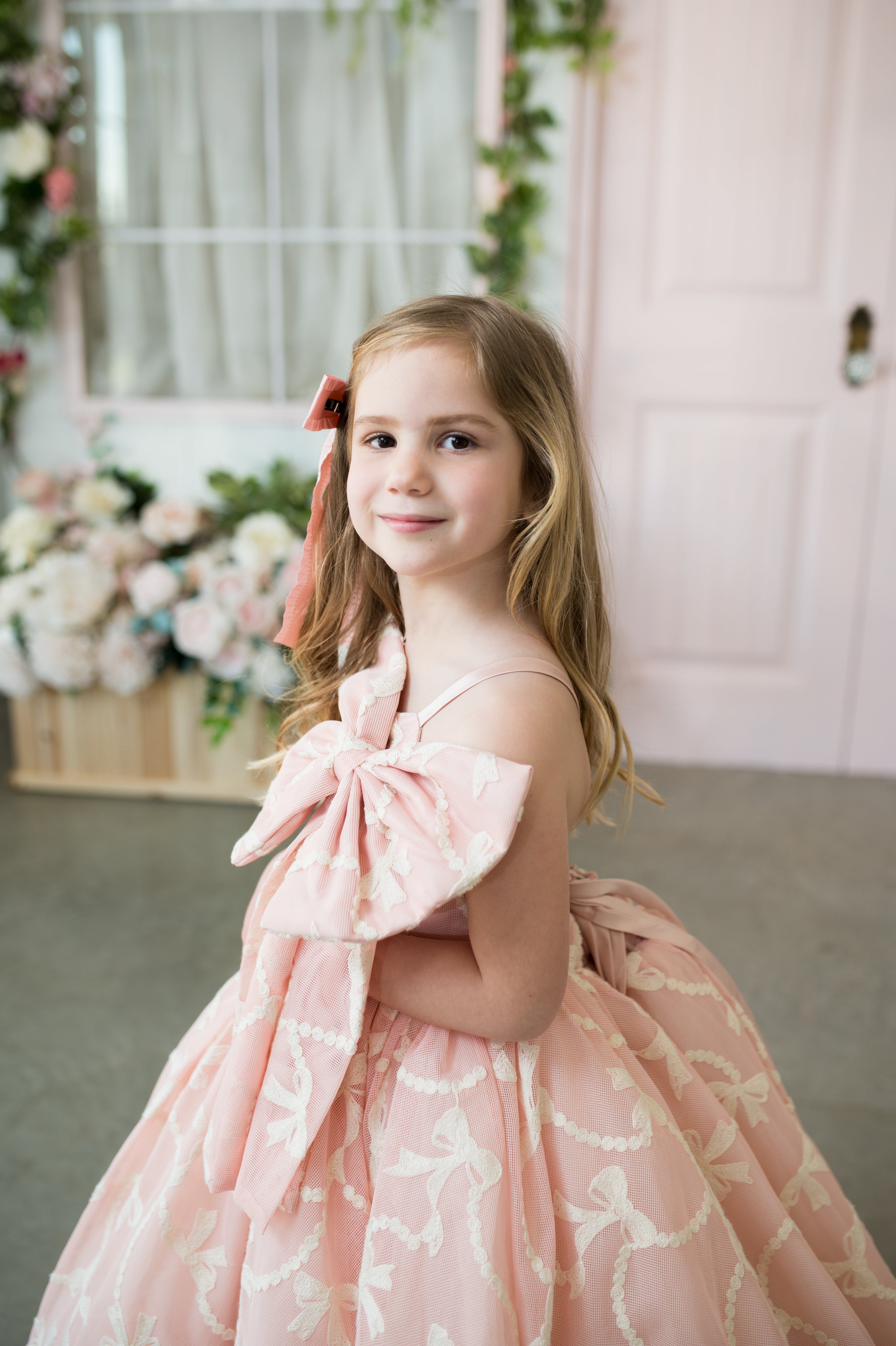 Young girl in a pink floral dress standing in a decorated room with flowers and a pink wall.