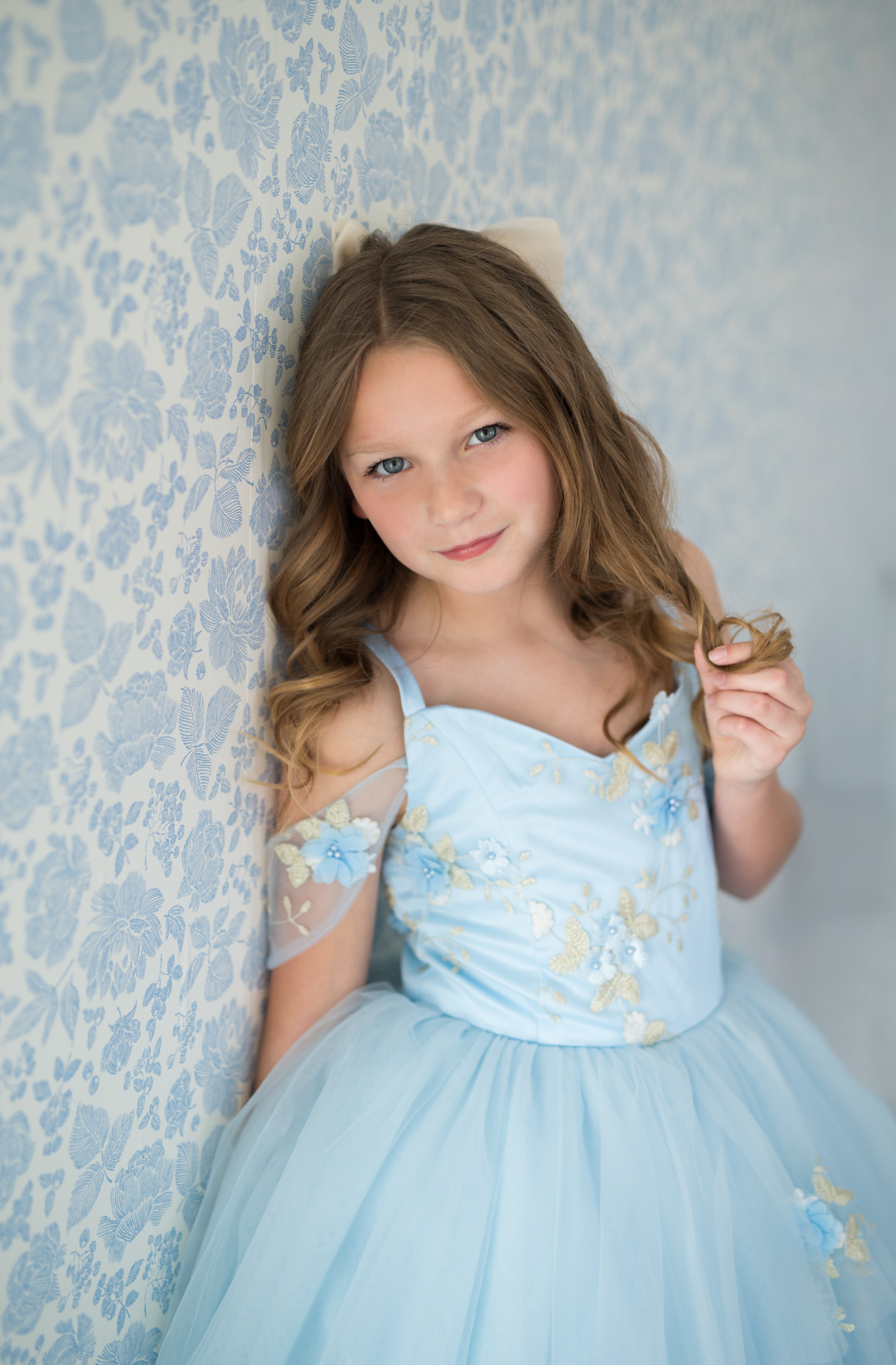 Young girl in a light blue floral dress standing against a floral wallpapered wall.