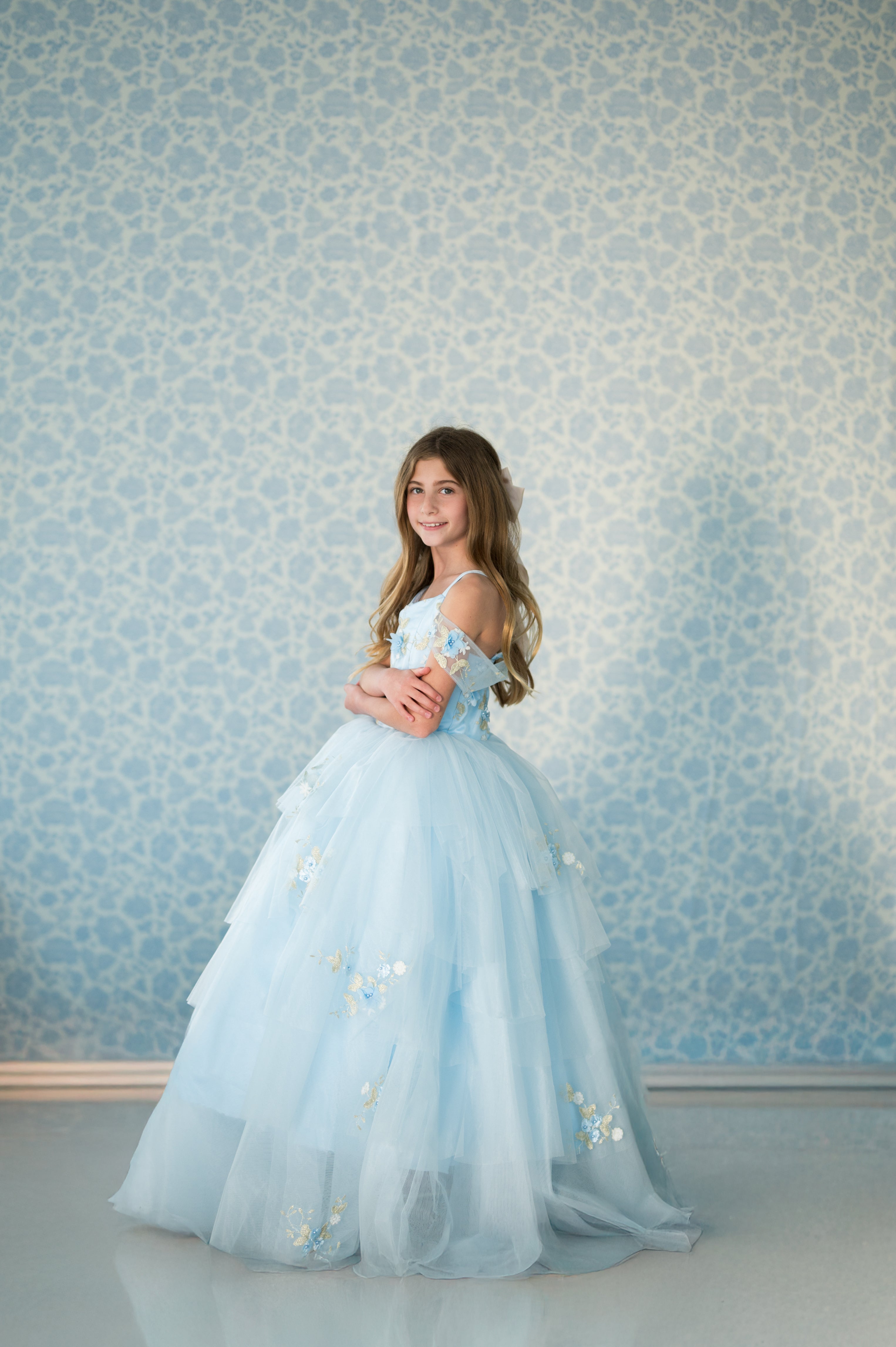 Young girl in a light blue dress standing against a patterned wall.