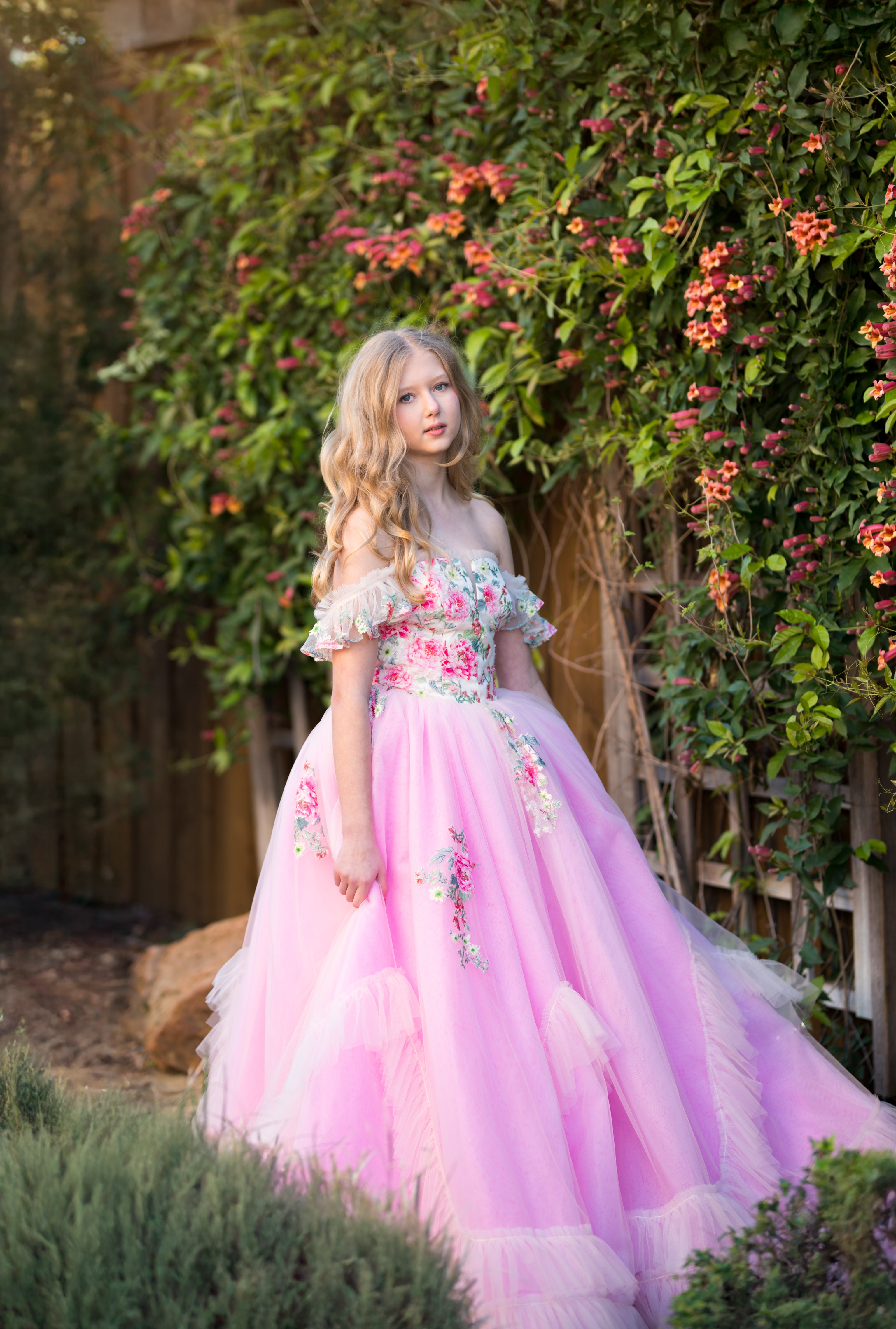 Young girl in a pink floral dress standing in a garden with flowers and greenery.