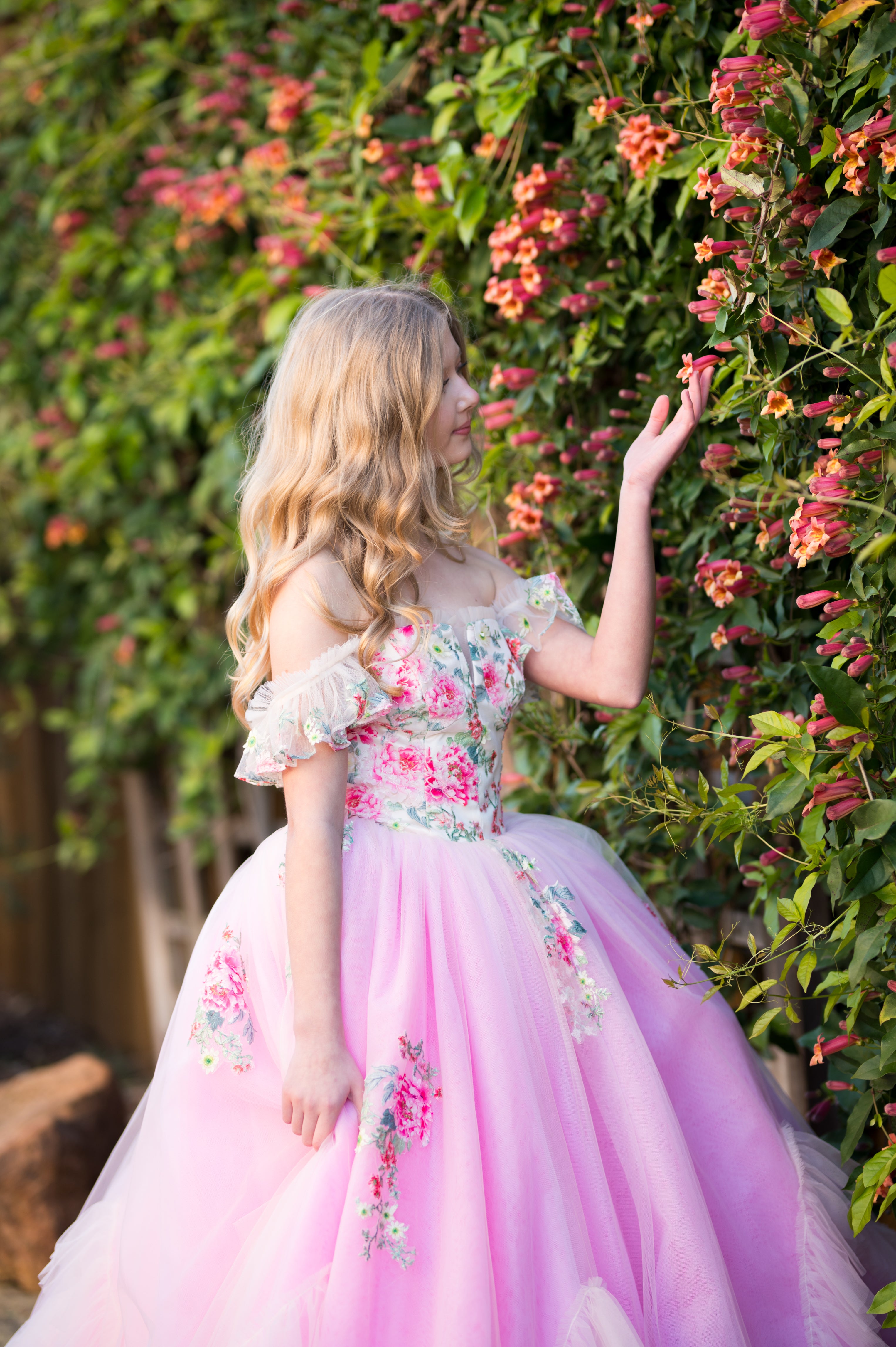Young girl in a pink floral dress standing in front of a flowering bush.