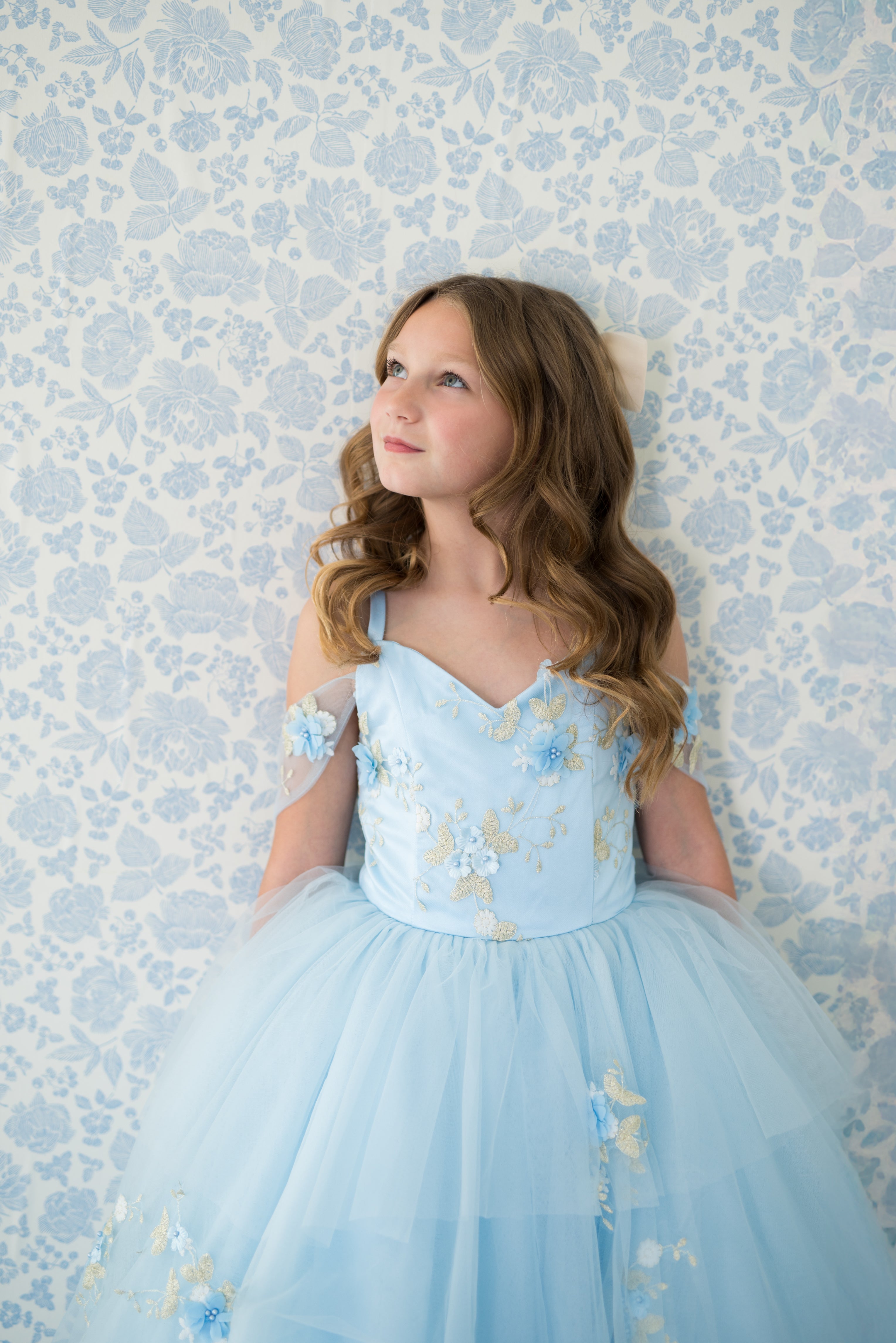 Young girl in a light blue dress with floral details against a floral wallpaper background