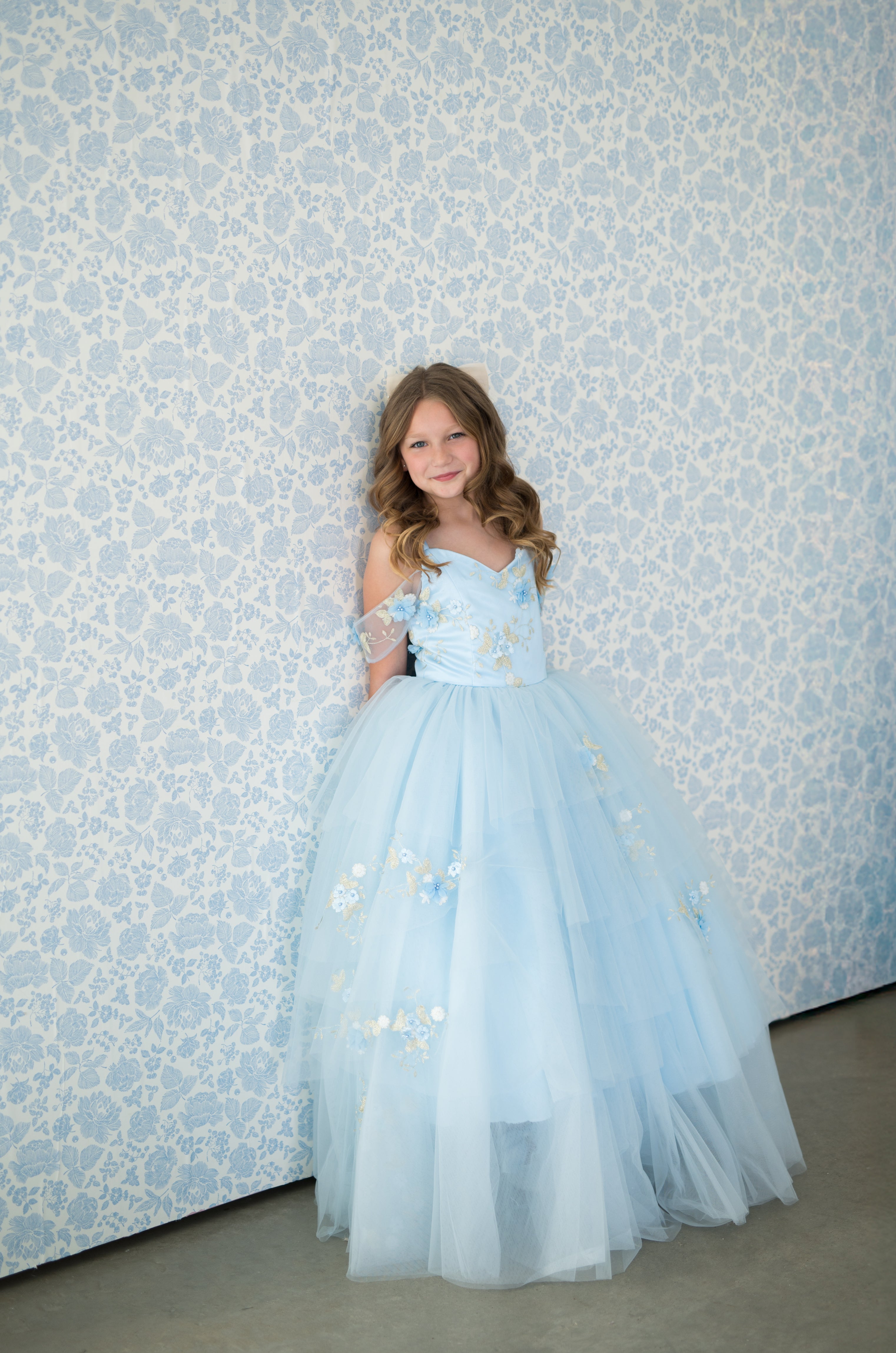 Young girl in a light blue dress standing against a patterned wall.