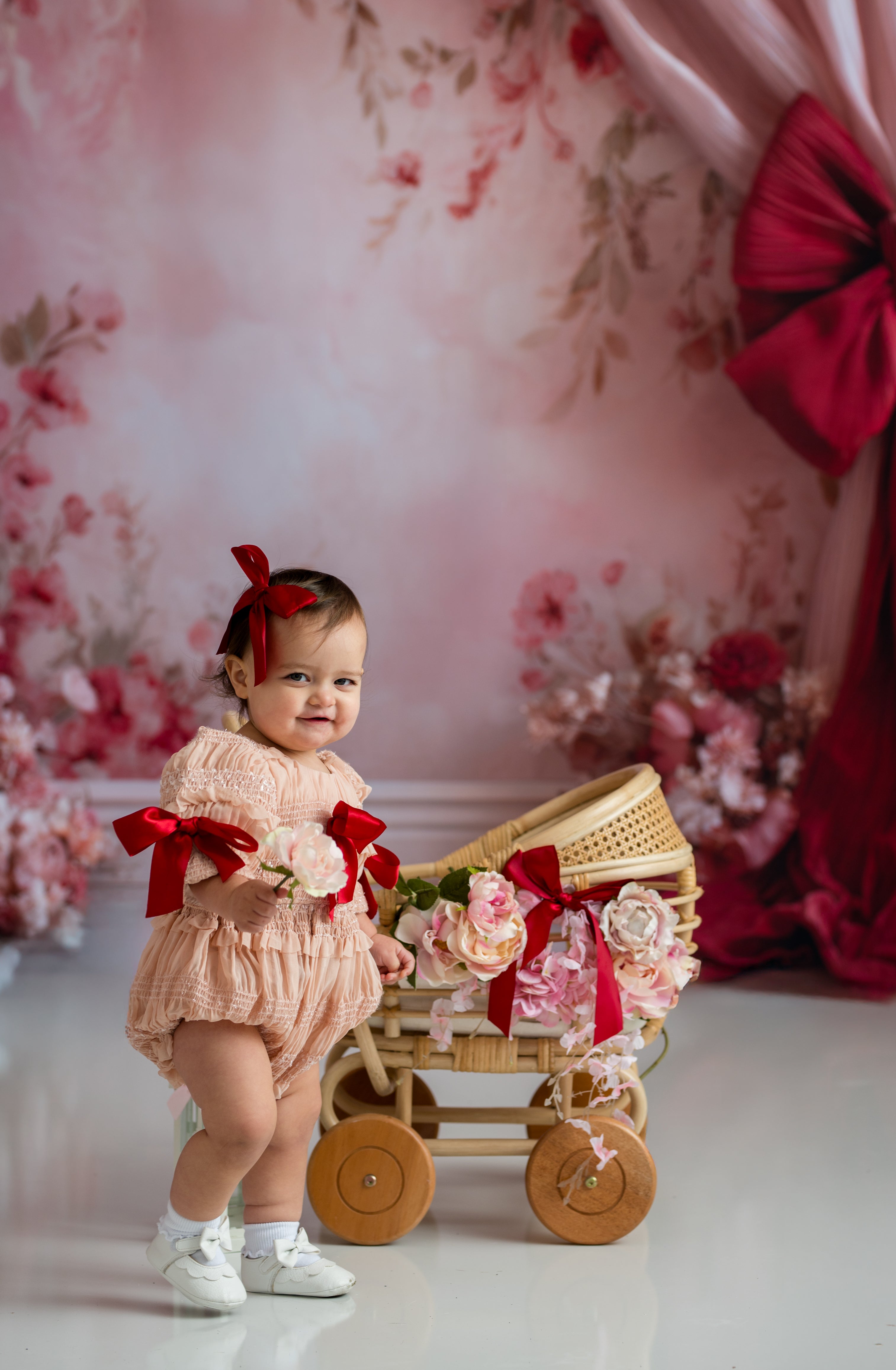 Baby in a pink outfit with a decorative cart against a floral backdrop