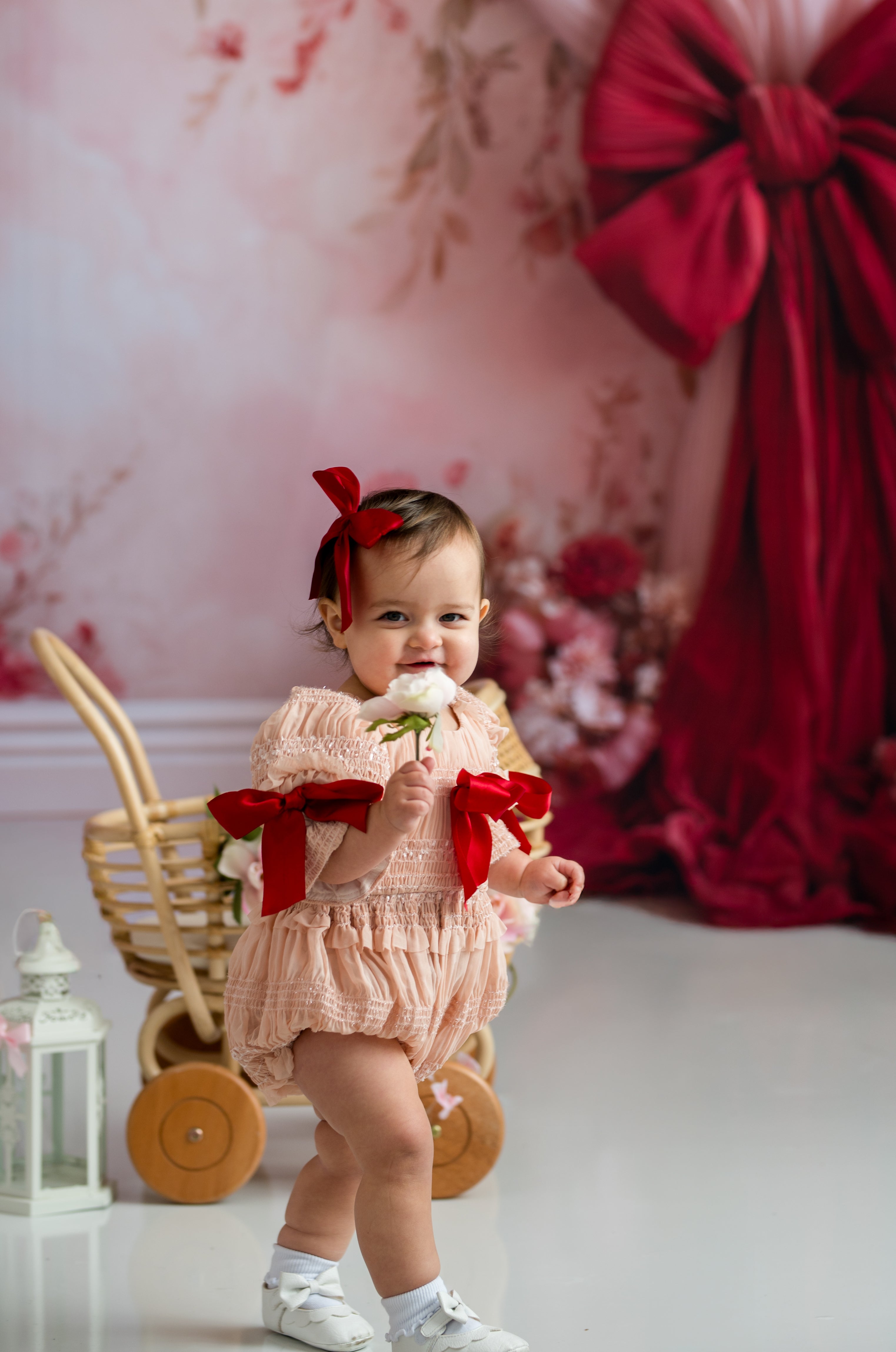 Baby in a pink outfit with red bows holding a flower, standing in front of a floral wall and wicker cart.