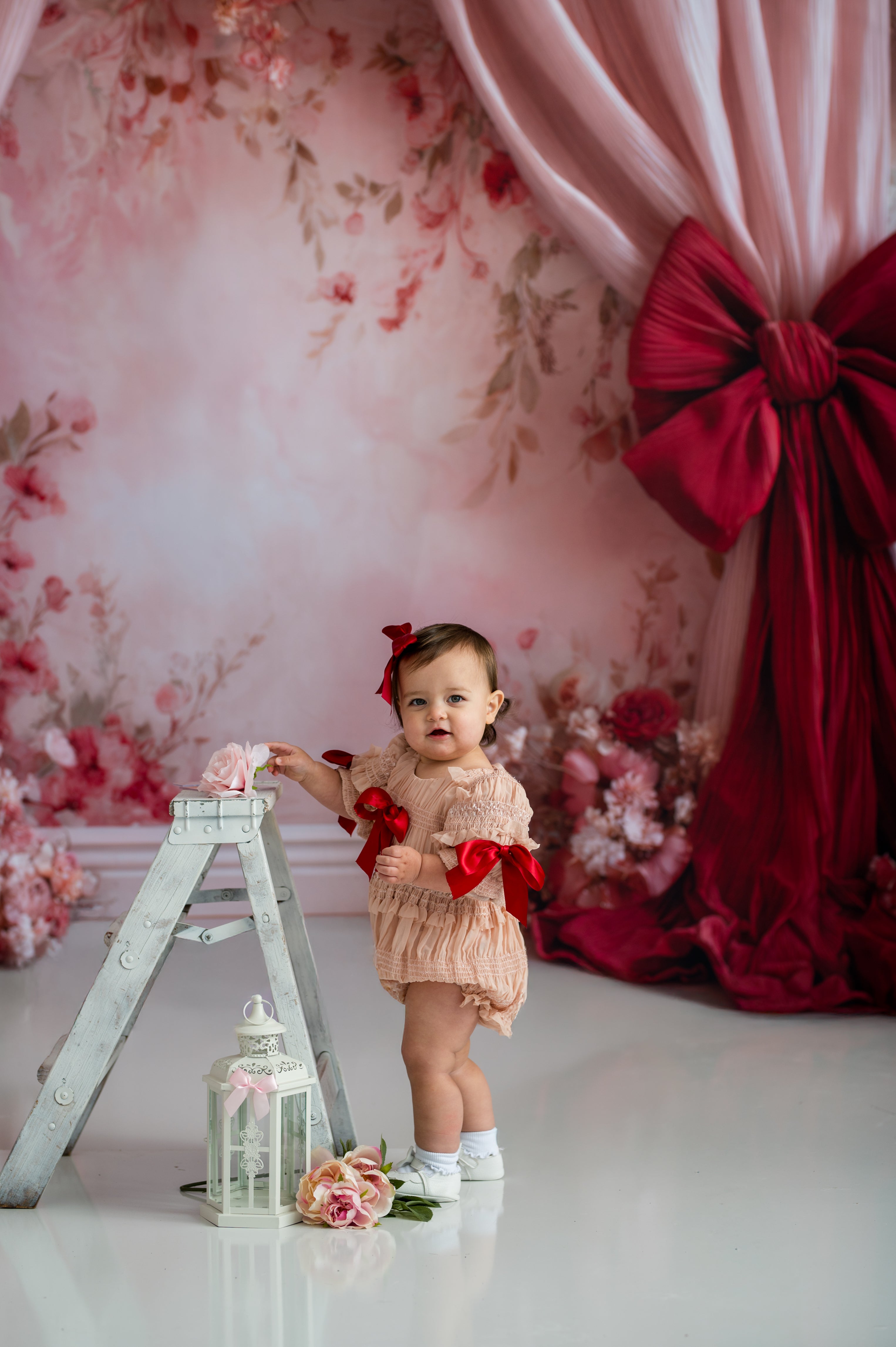 Baby in a lace outfit with red bows standing next to a decorative ladder against a floral and red curtain backdrop.