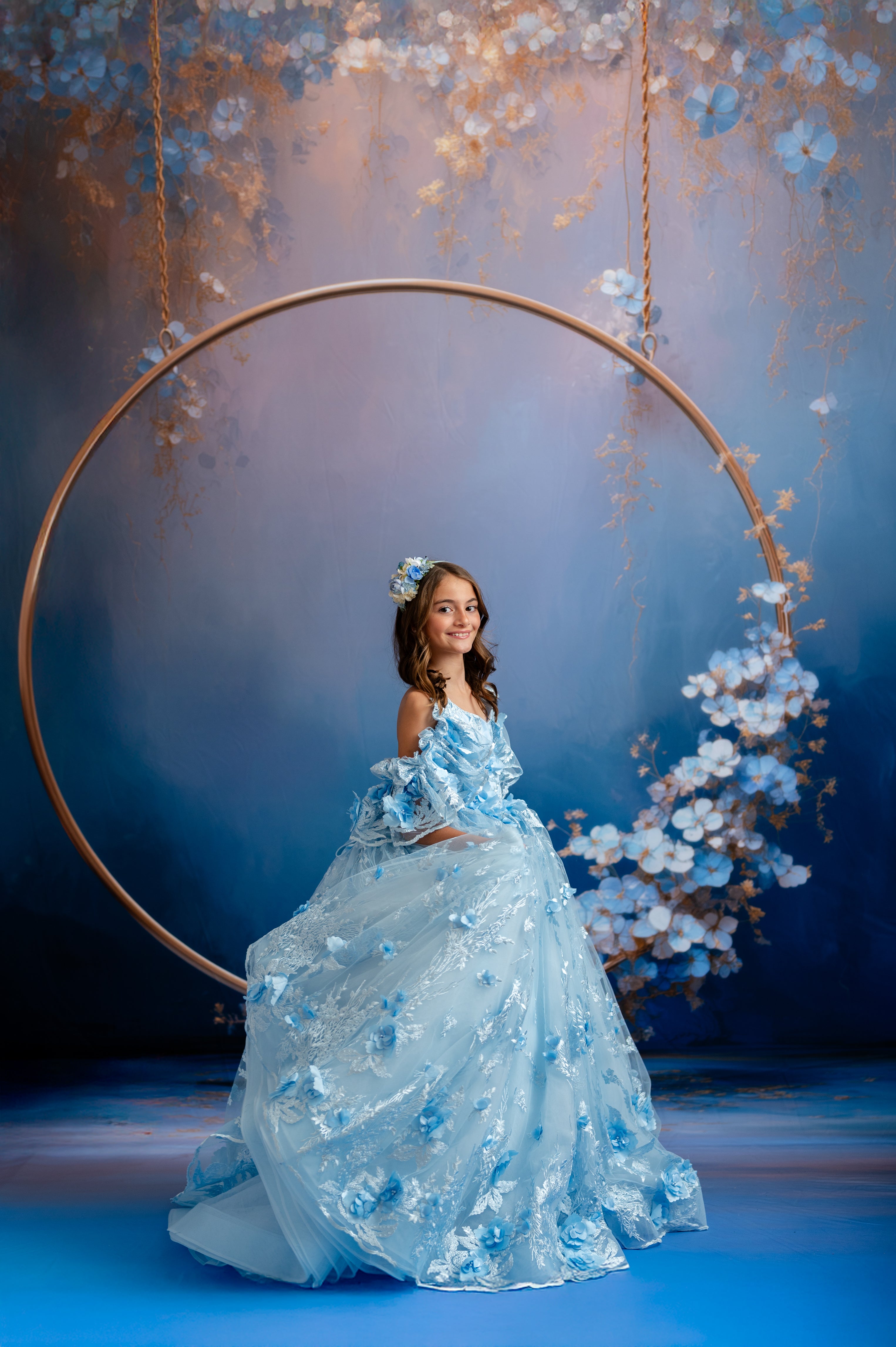 Woman in a blue floral dress sitting on a circular swing with floral decorations against a dark background