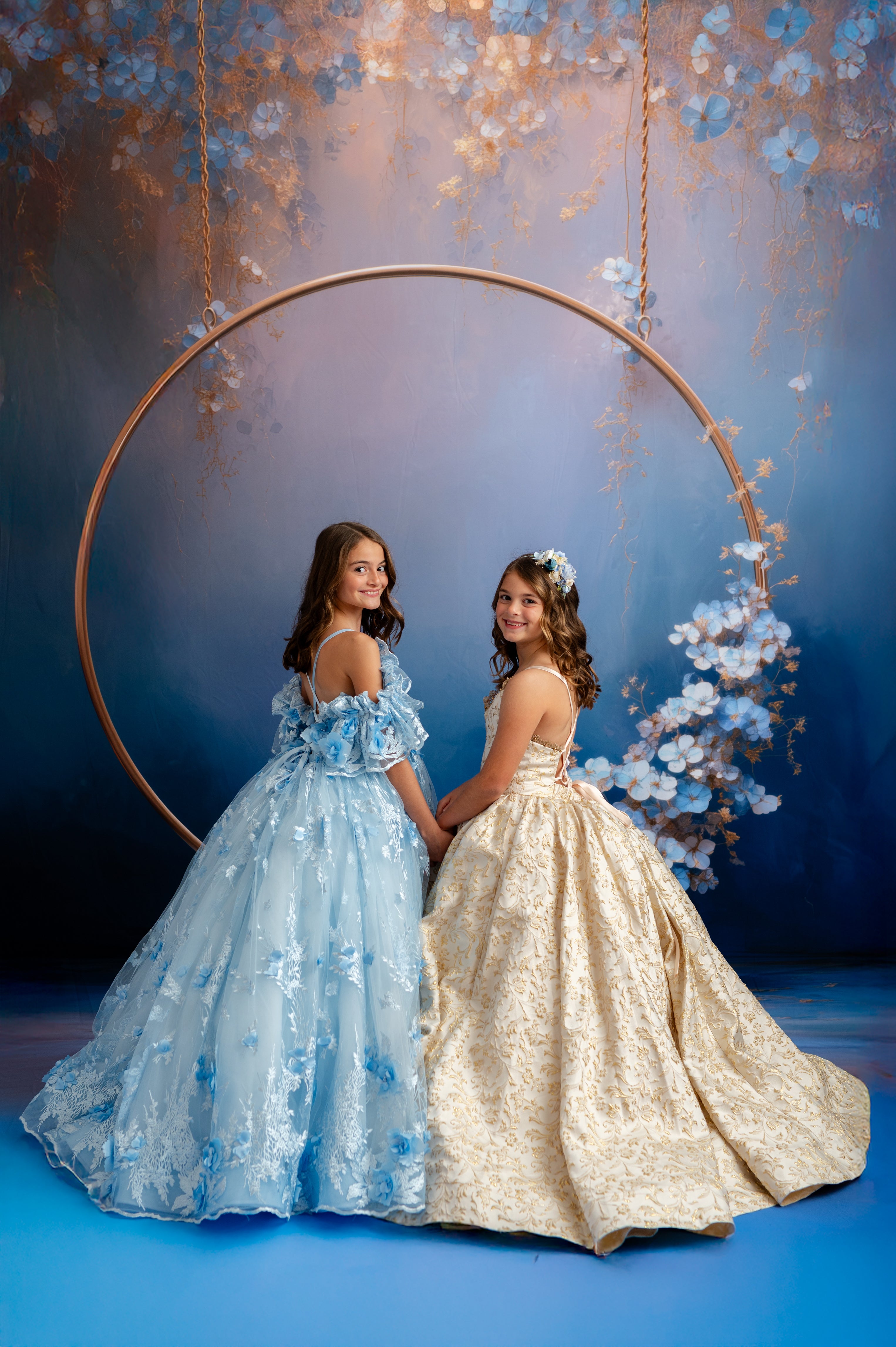 Two women in elegant dresses standing next to a floral hoop against a blue background