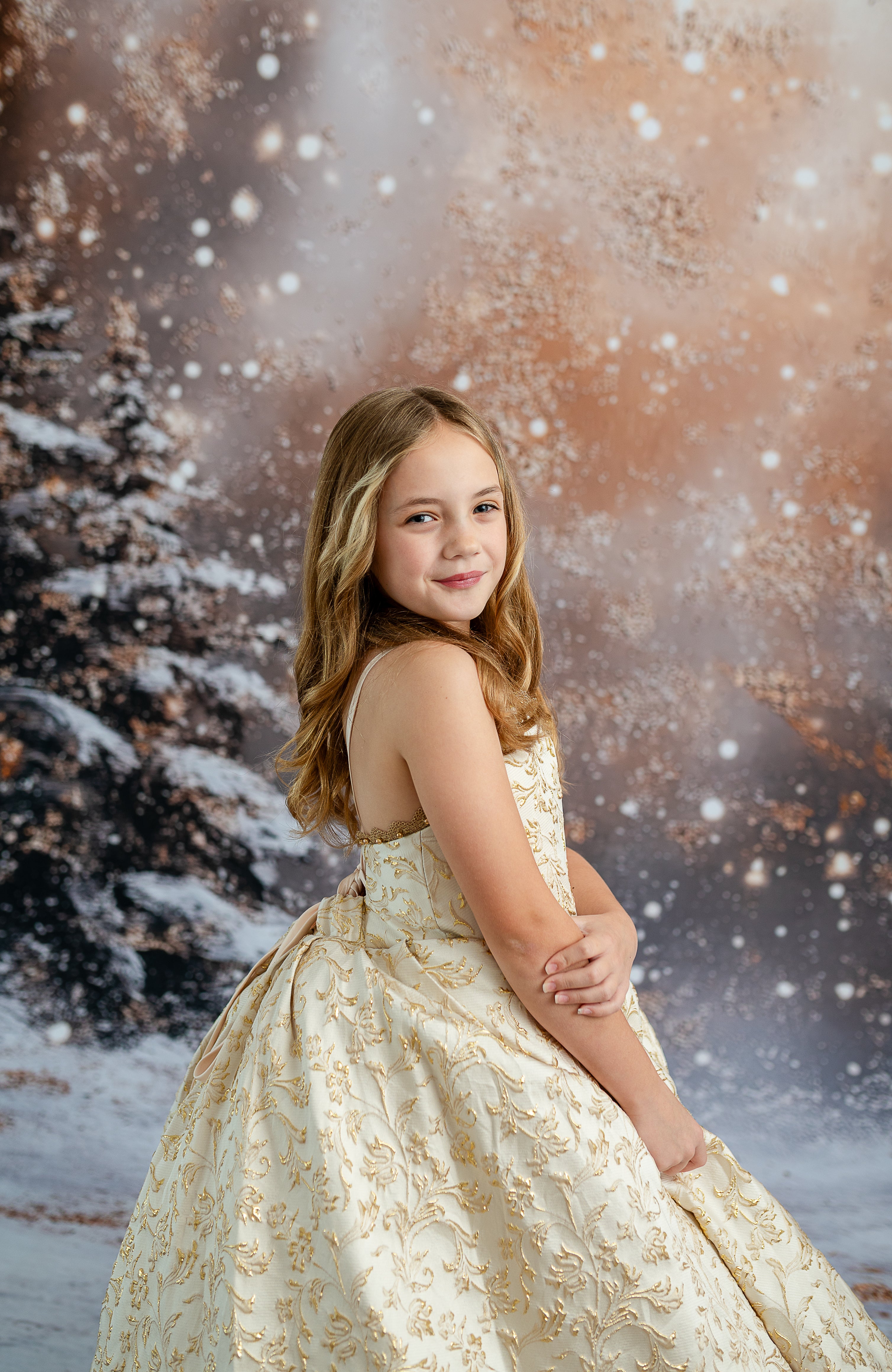 Young girl in a gold dress standing in front of a snowy tree backdrop