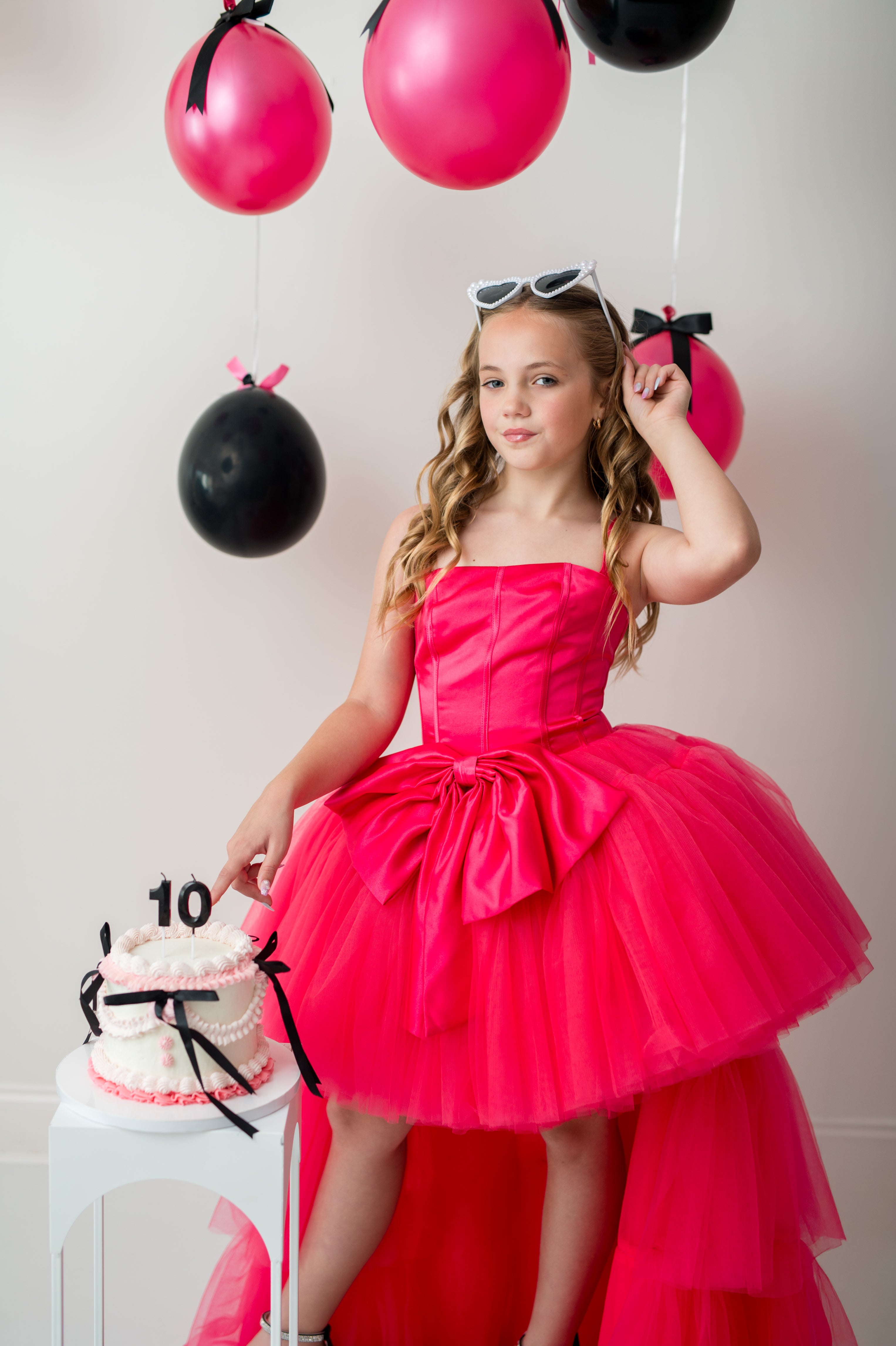 Young girl in a pink dress standing next to a birthday cake with balloons in the background.