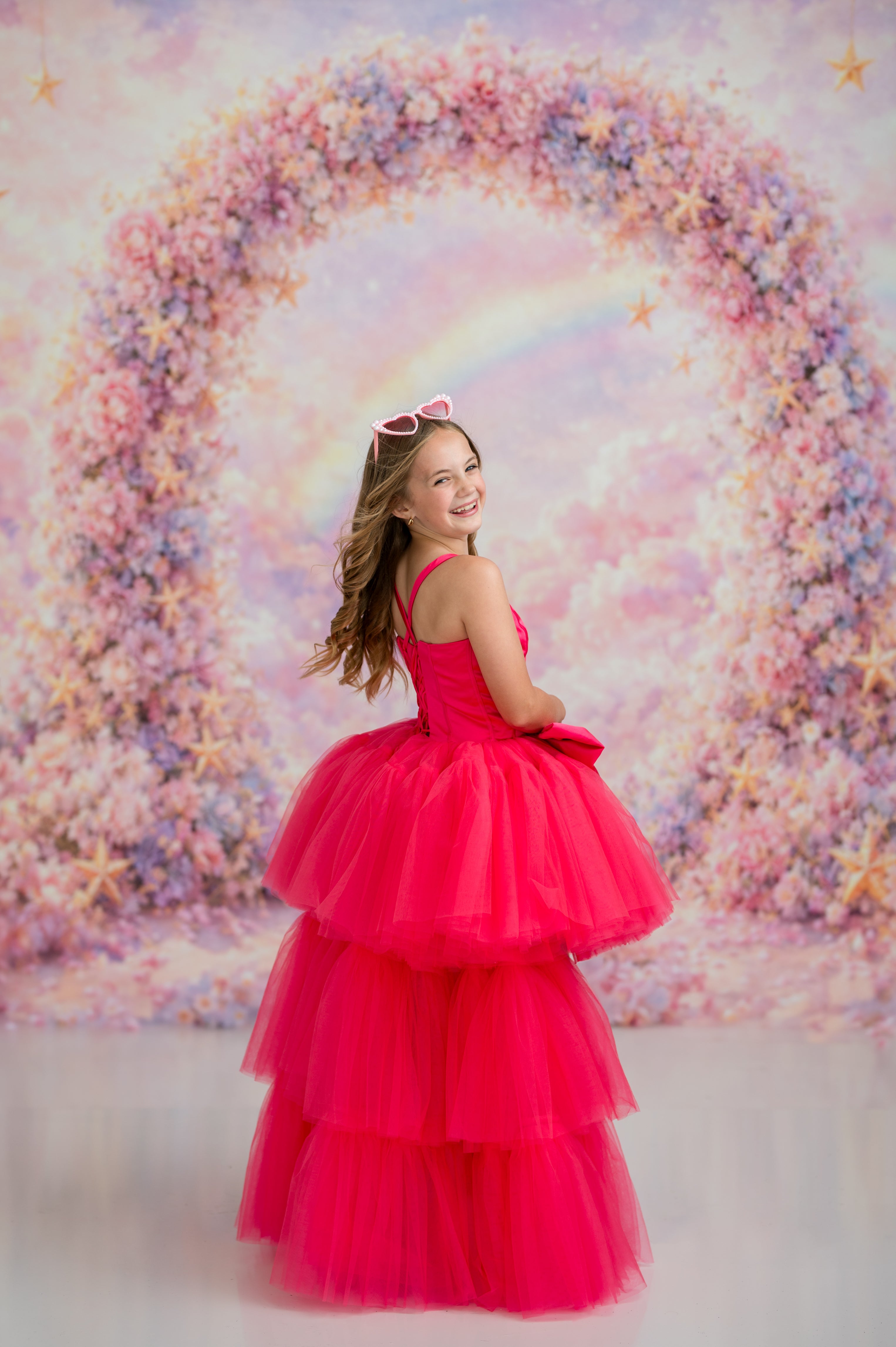 Young girl in a pink dress standing in front of a colorful floral arch with a rainbow.