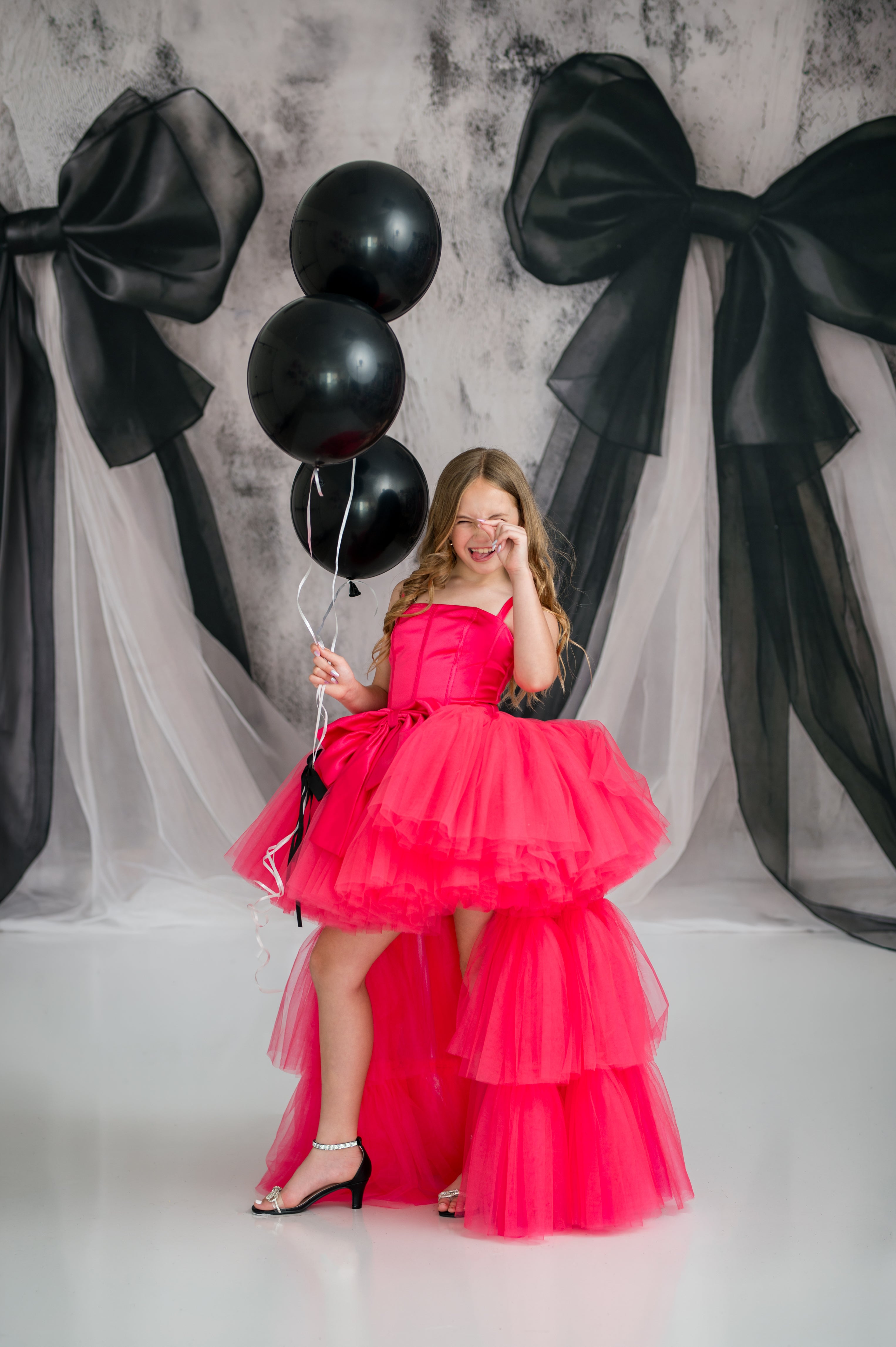Young girl in a pink dress holding black balloons against a backdrop with large black bows.