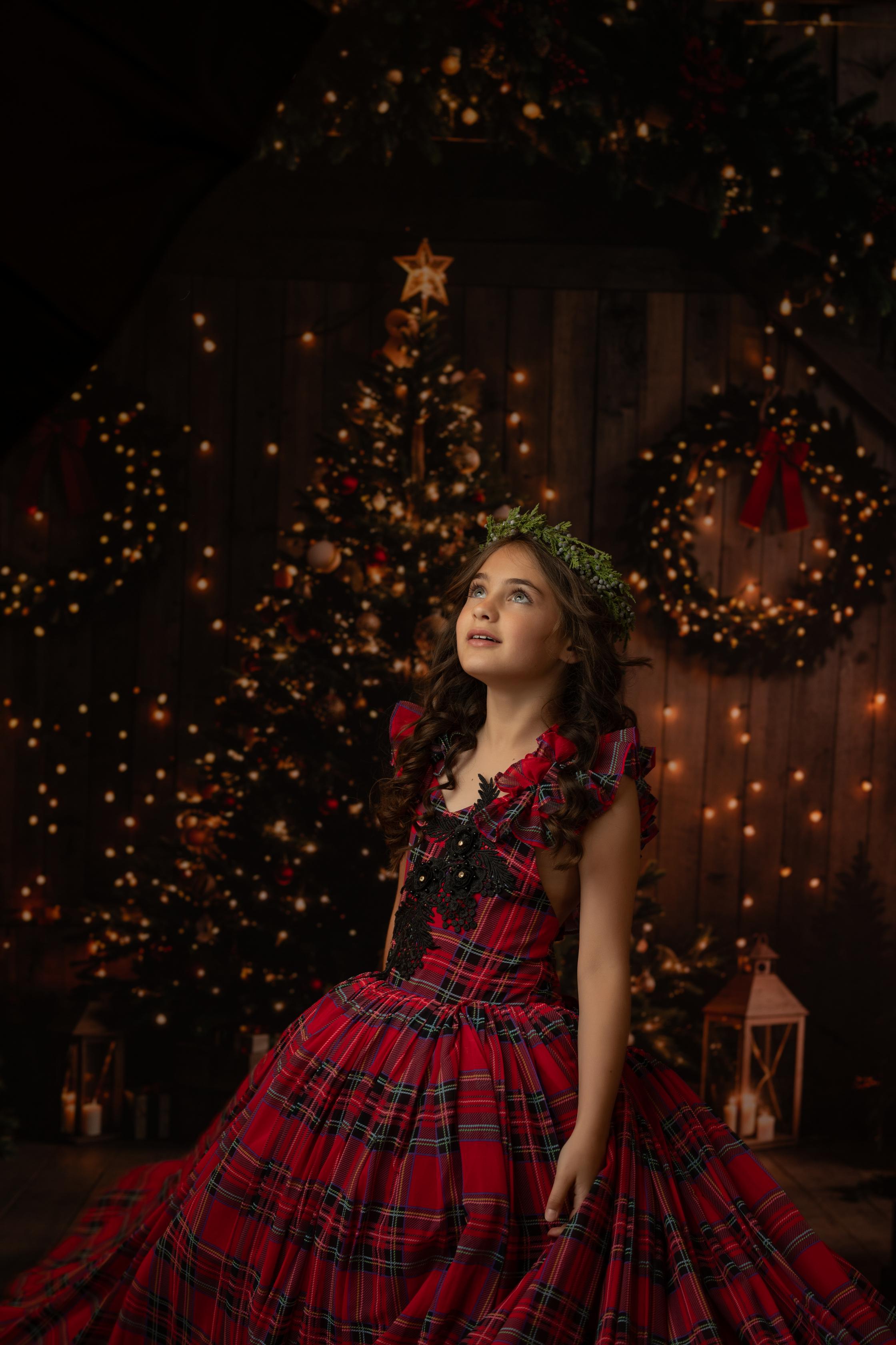 Little girl posing in a red and green plaid gown, with twinkling Christmas lights and decorations behind her.