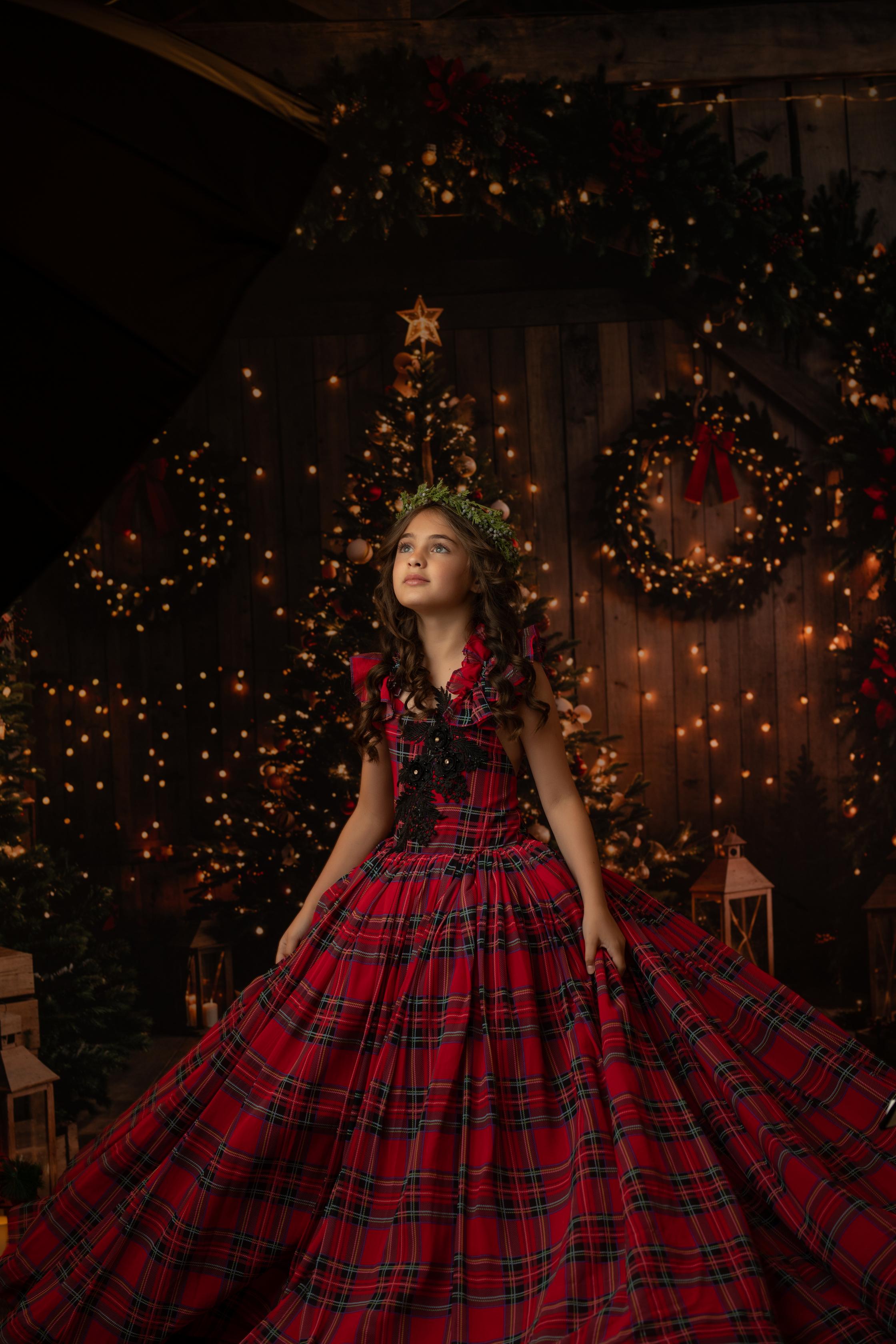 A girl dressed in a red and black tartan dress with a black floral lace accent, standing in front of a warmly lit Christmas tree.