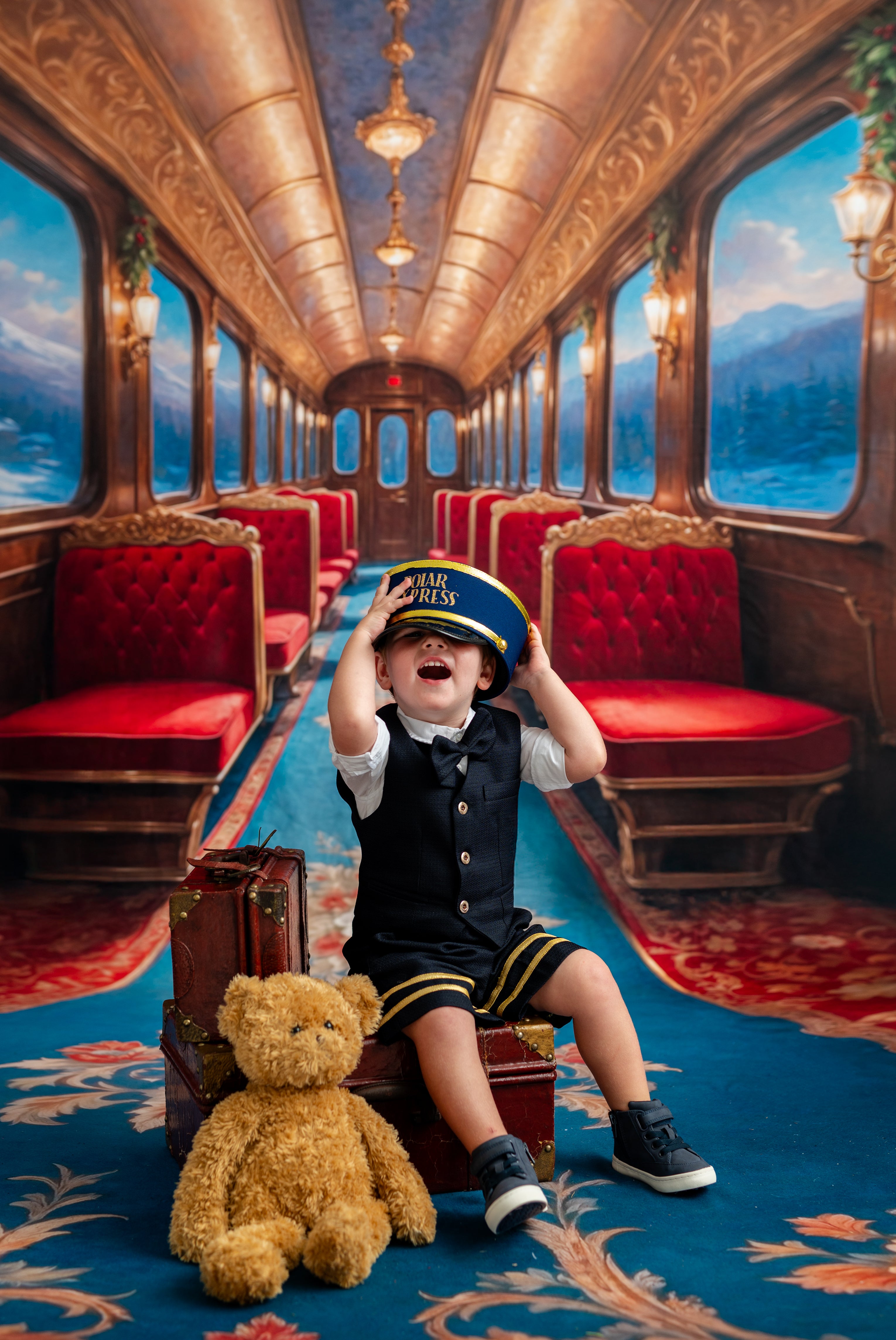 Child in a train car with red seats and a teddy bear, wearing a pilot outfit.