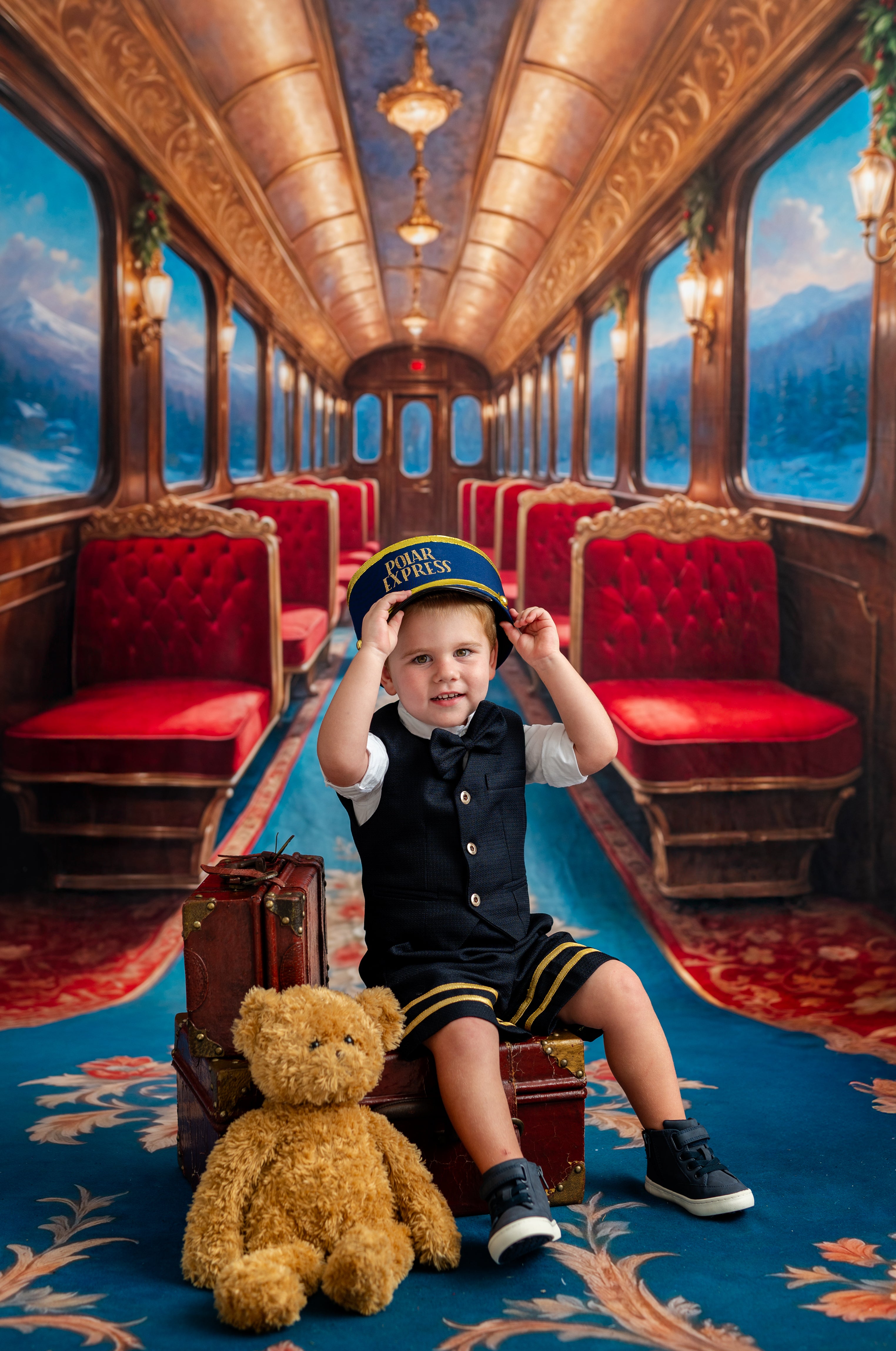 Child in a train car with red seats and a teddy bear, wearing a uniform and hat.