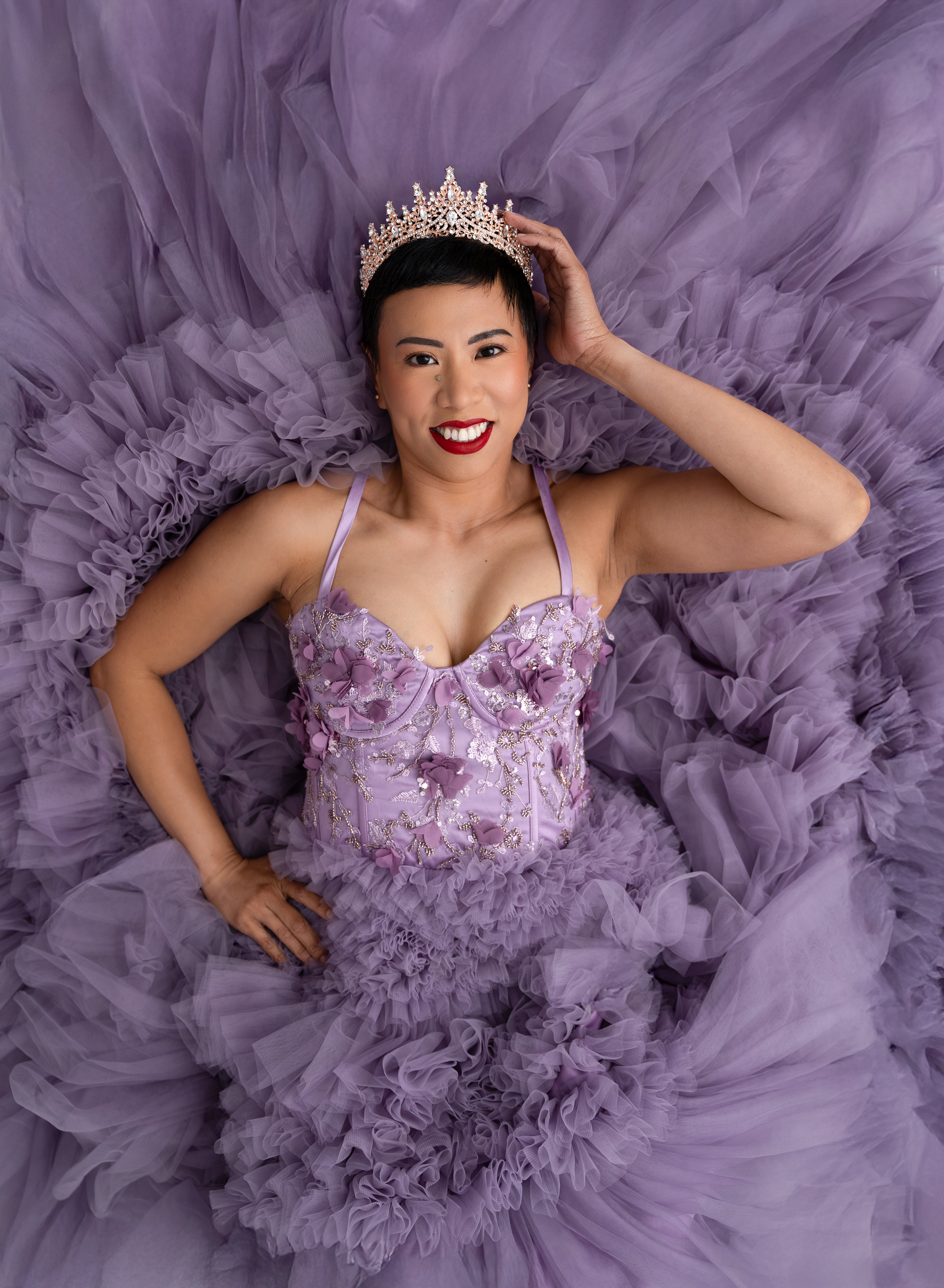 Woman in royal-themed attire, wearing a sparkly crown and lavender gown with playful expression in a decorative setting.