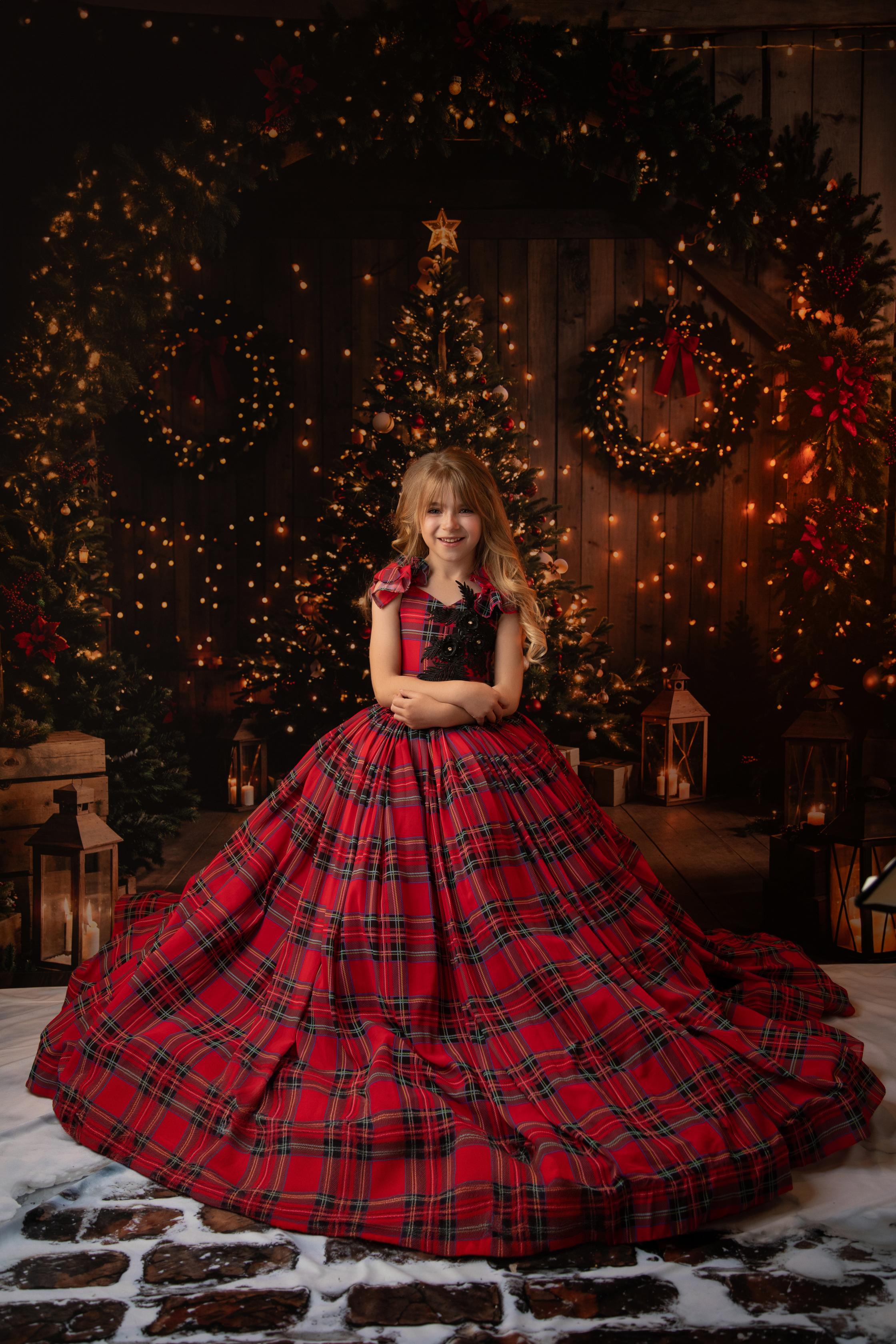 A young girl twirling in a red plaid gown adorned with black lace details, with a warmly lit Christmas tree behind her.
