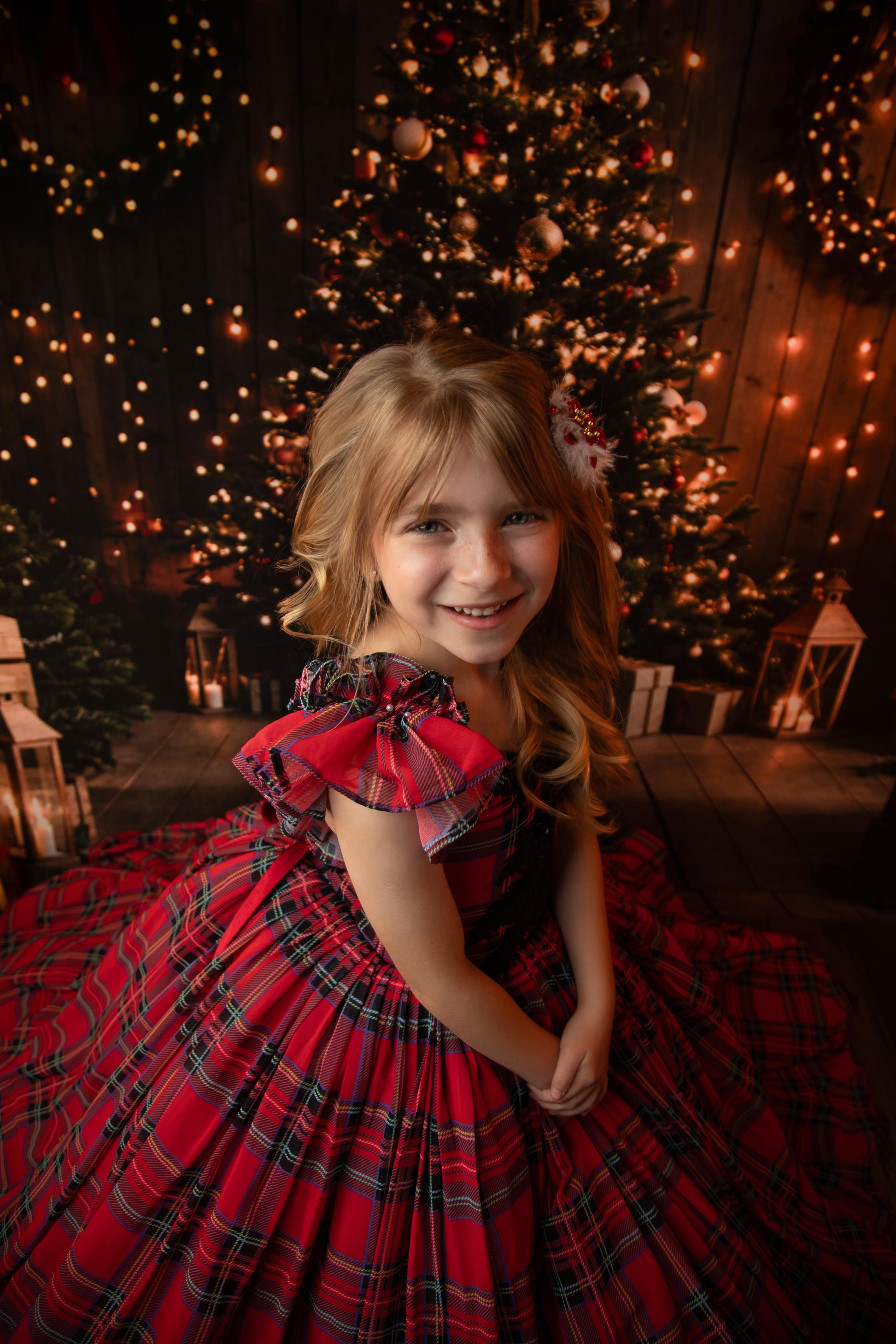 Child in a red tartan holiday dress, with intricate black lace detailing, posing in front of a decorated Christmas tree.
