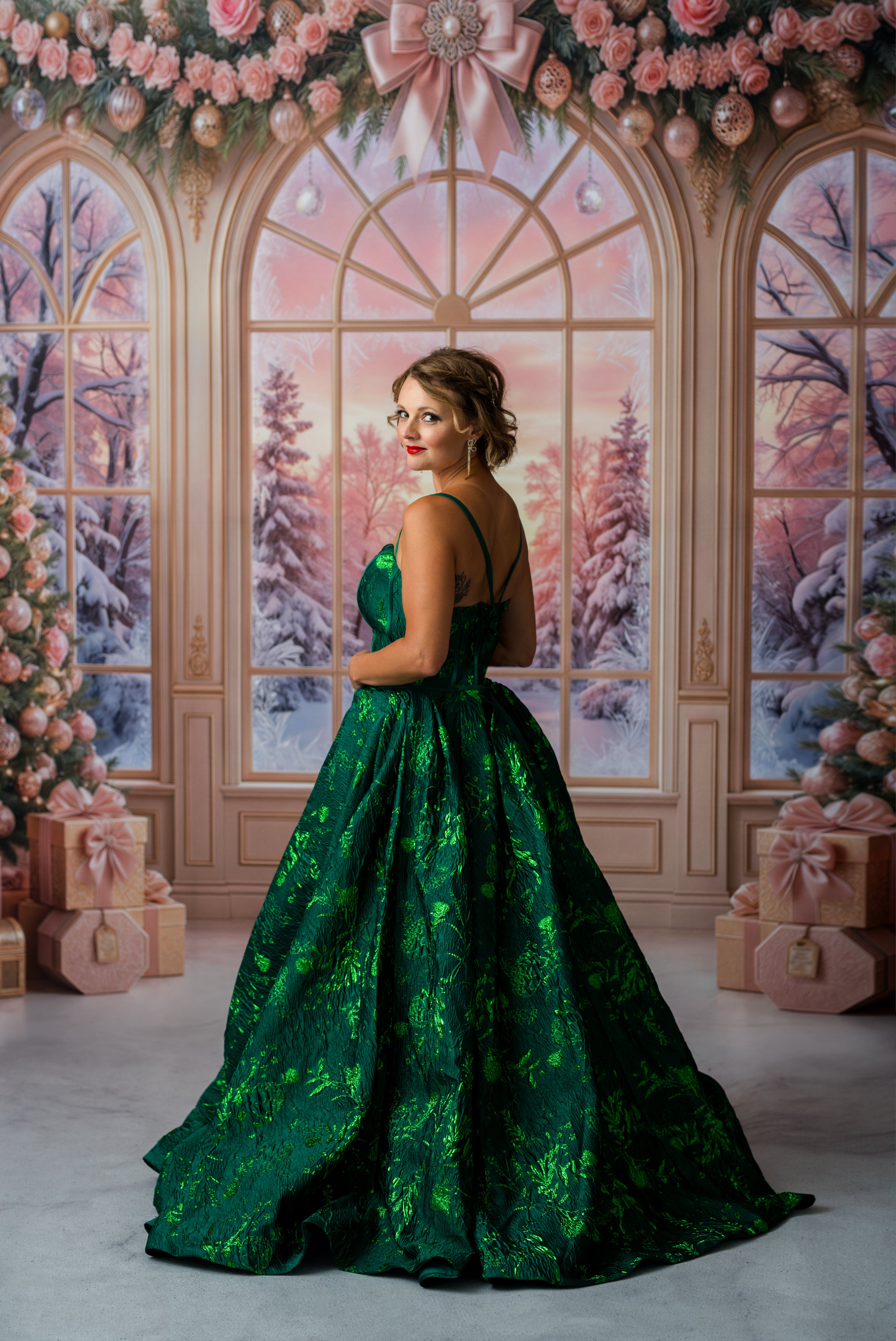 A woman and two young girls pose together in an ornate holiday-themed room. The woman wears a floor-length emerald green gown, and the two girls wear matching green dresses with different skirt details, one with a red tulle overlay and one with a yellow tulle overlay.