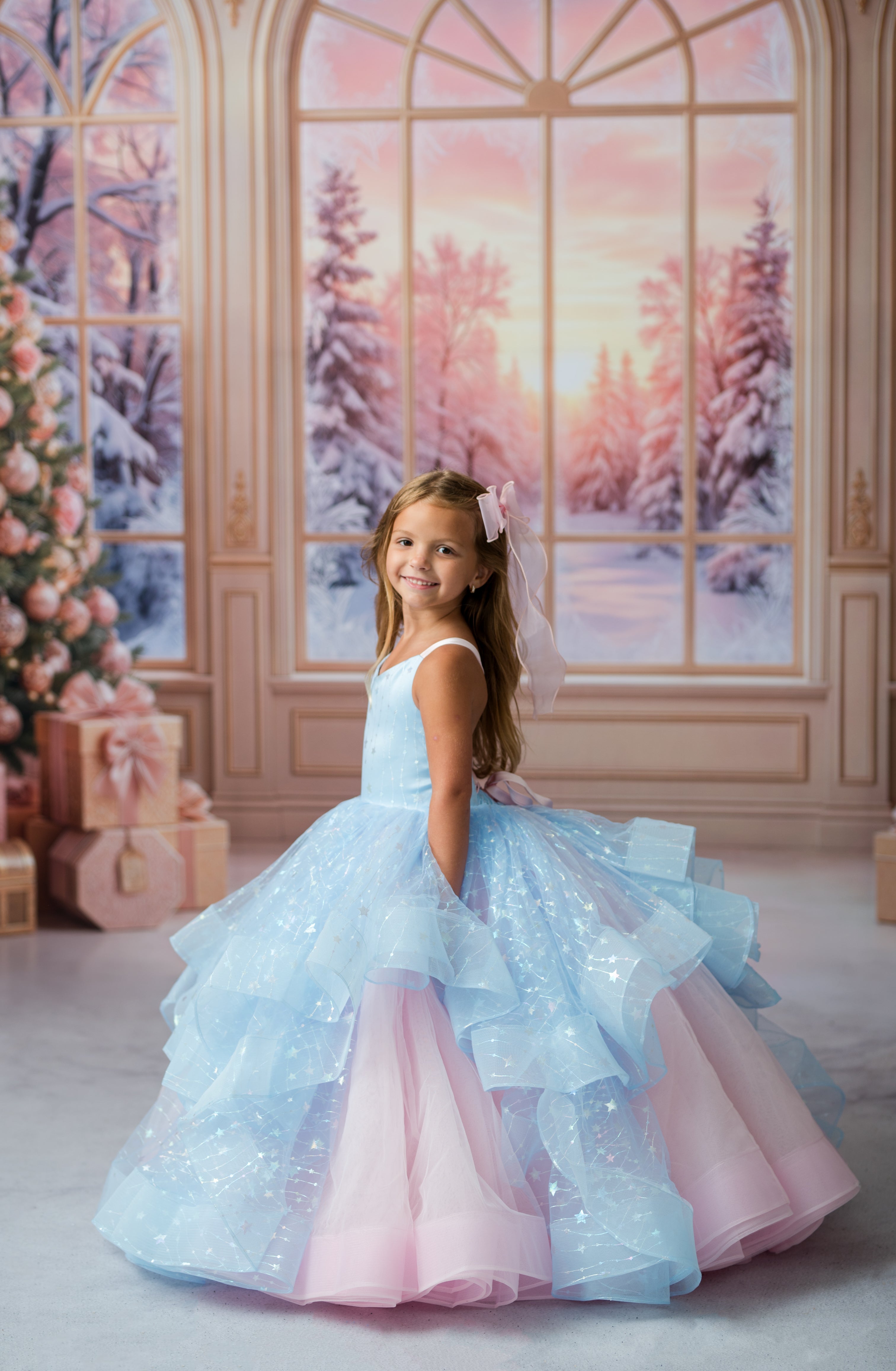 A young girl with long light brown hair wearing a voluminous light blue and pink ball gown with a sheer veil, standing in front of a festive holiday backdrop with large windows showing a snowy winter forest scene.
