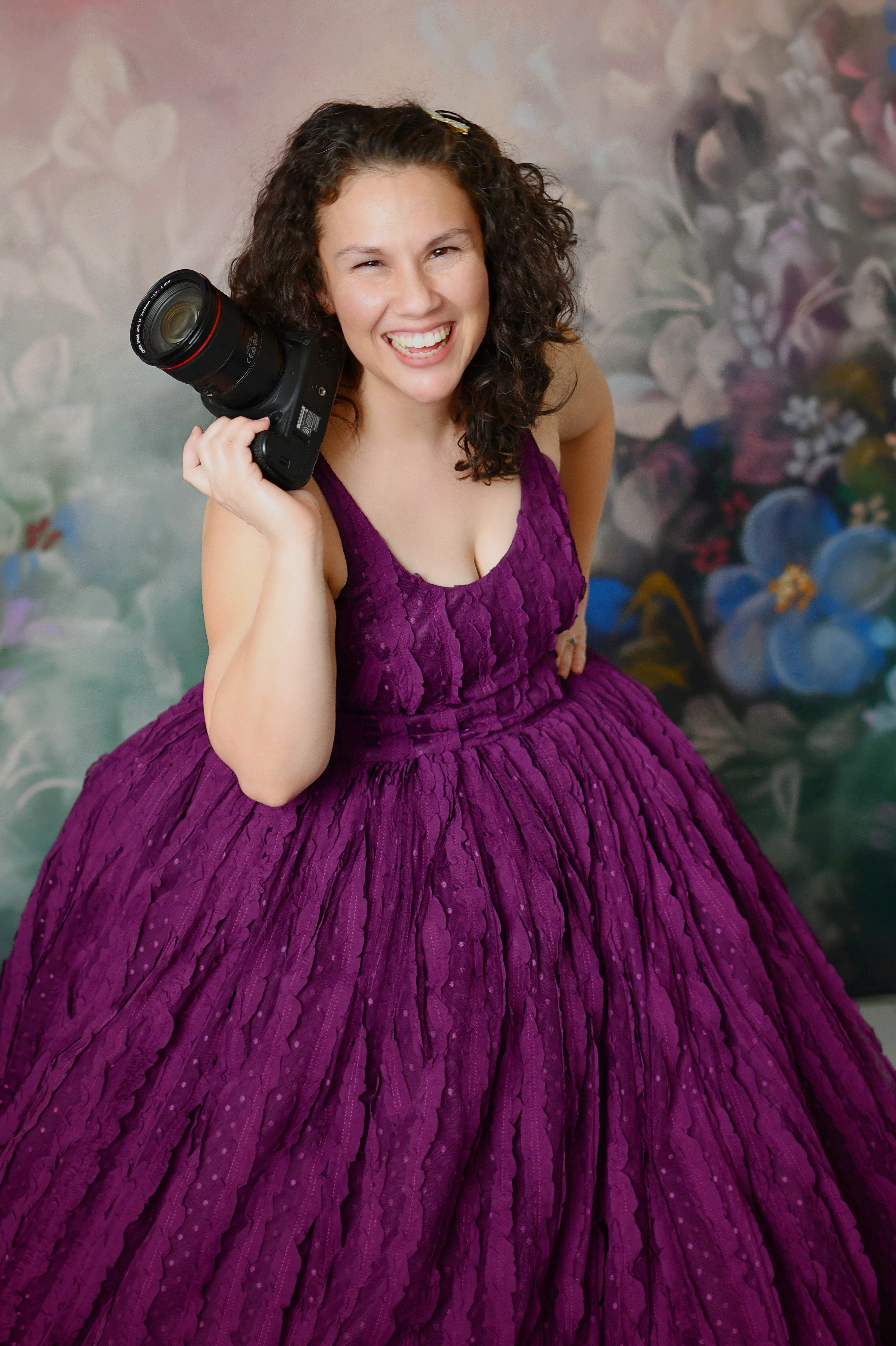 A woman in a floor-length, solid purple gown with a voluminous skirt, smiling and holding a camera.