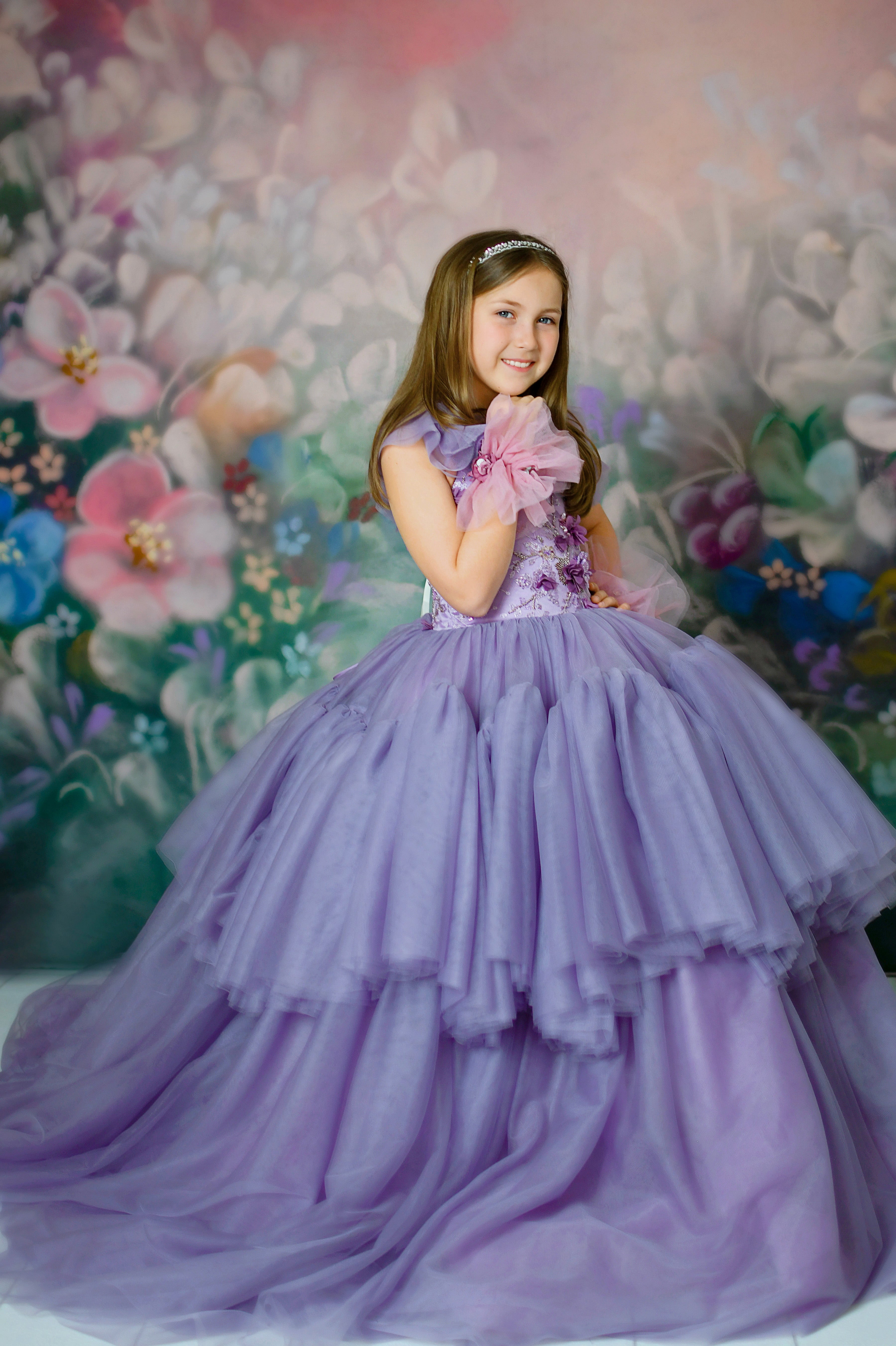 "Young girl in a luxurious violet floor-length gown with floral appliqués, tiered tulle skirt, and sheer ruffle details, posing against a dreamy floral backdrop."