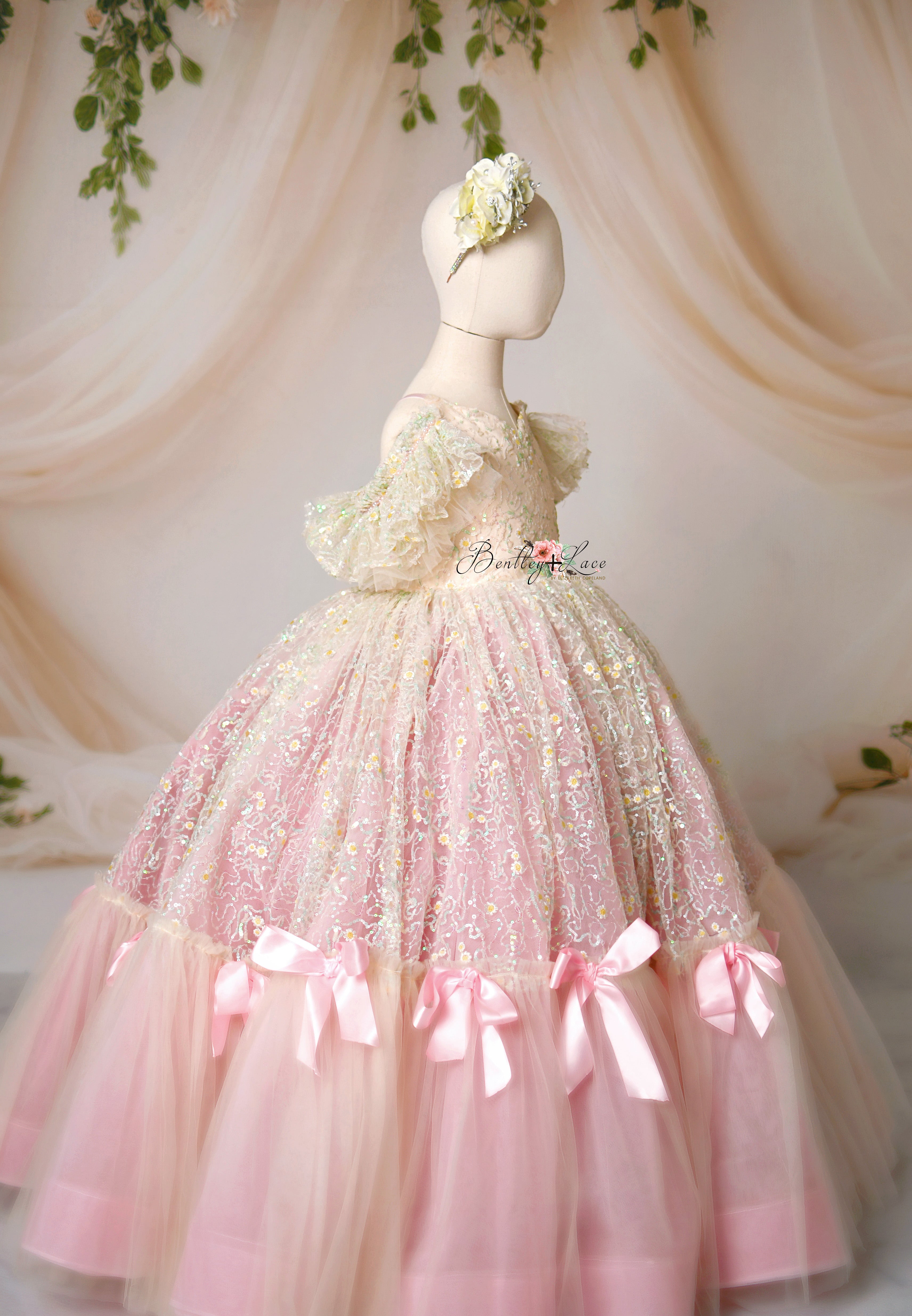 A floor-length pink gown with daisy sequin-embroidered lace and light pink bows, displayed on a mannequin.