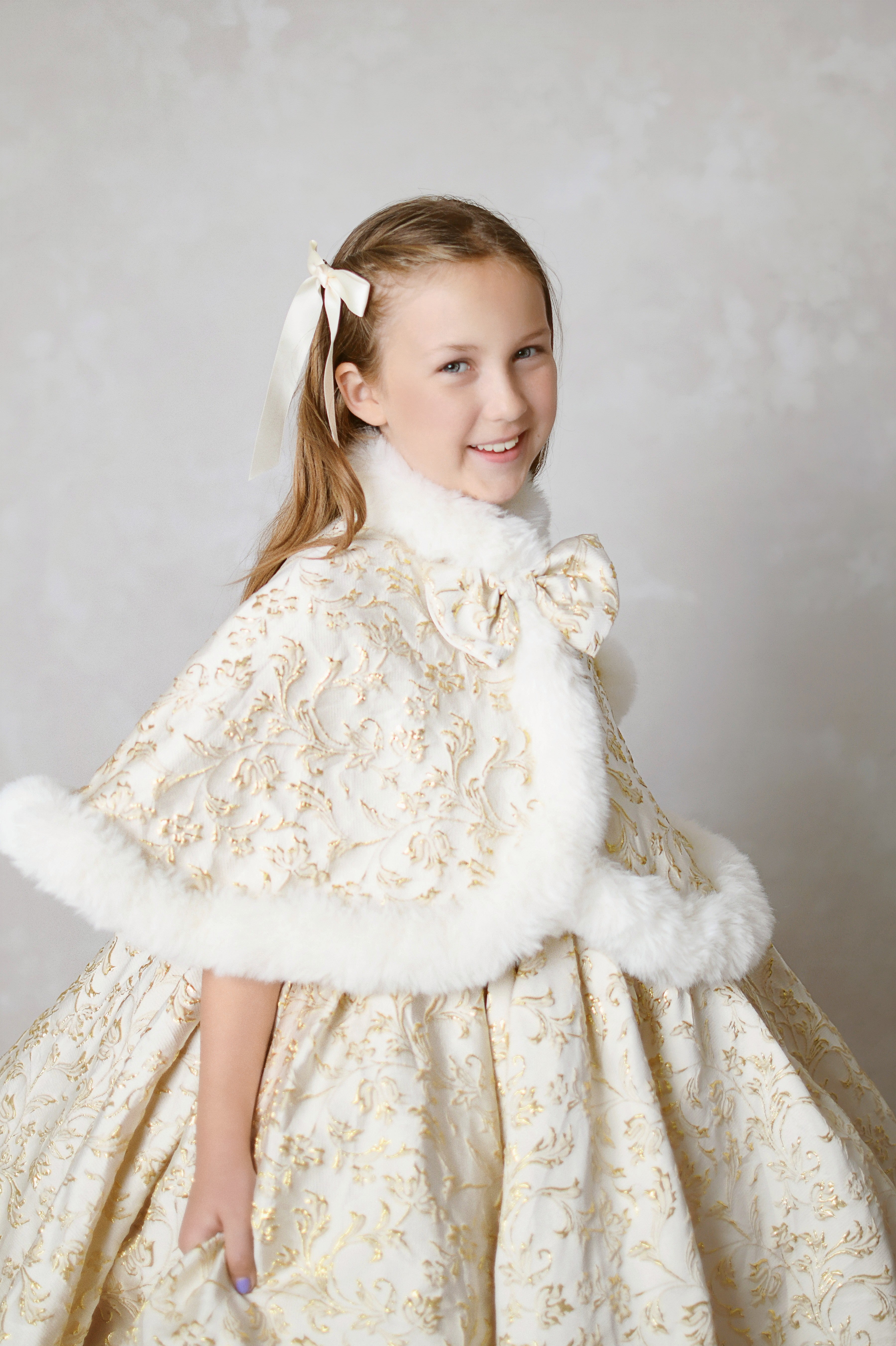 Child model wearing an elegant ivory and gold ball gown with a full skirt, standing in a holiday-themed photography setting.