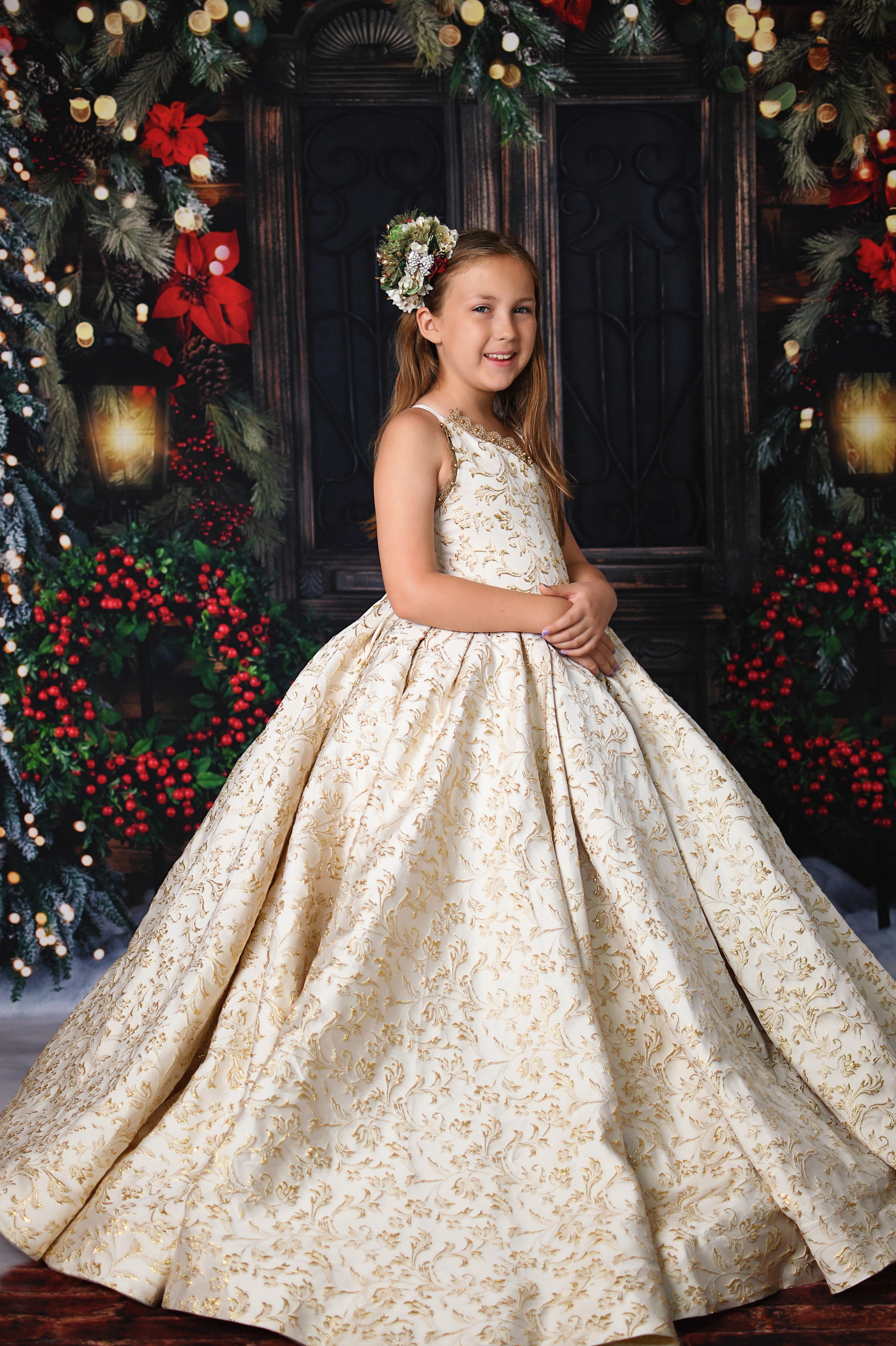 Young girl in a gold brocade holiday gown posing in front of a festive Christmas backdrop with pine garlands and red poinsettias.