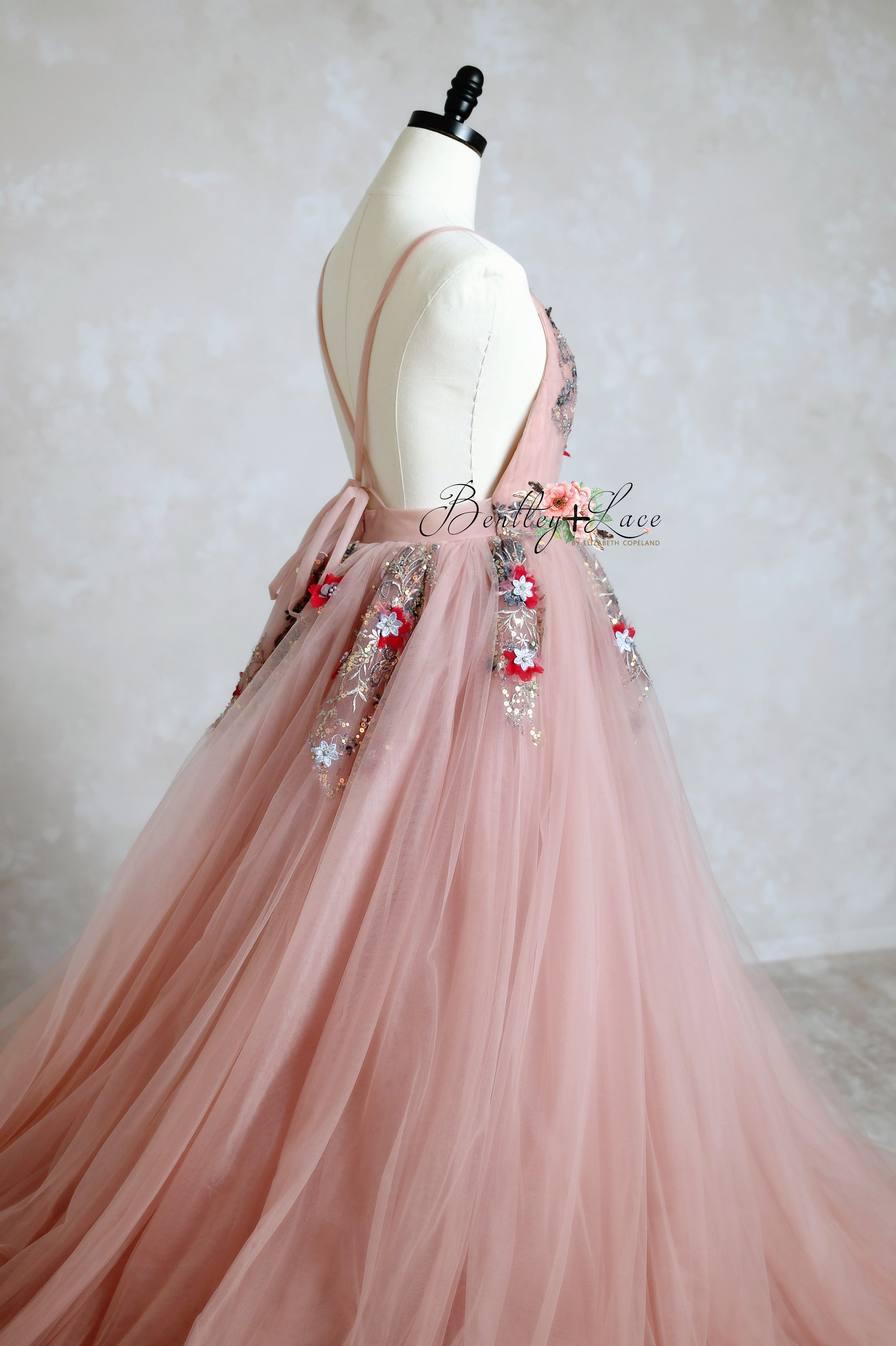 The bohemian Dillon gown features a charming silhouette in rose blush, with a deep V-cut bodice, delicate pleating, hand-sewn floral appliques, an open back, and a layered silk tulle skirt with a train.