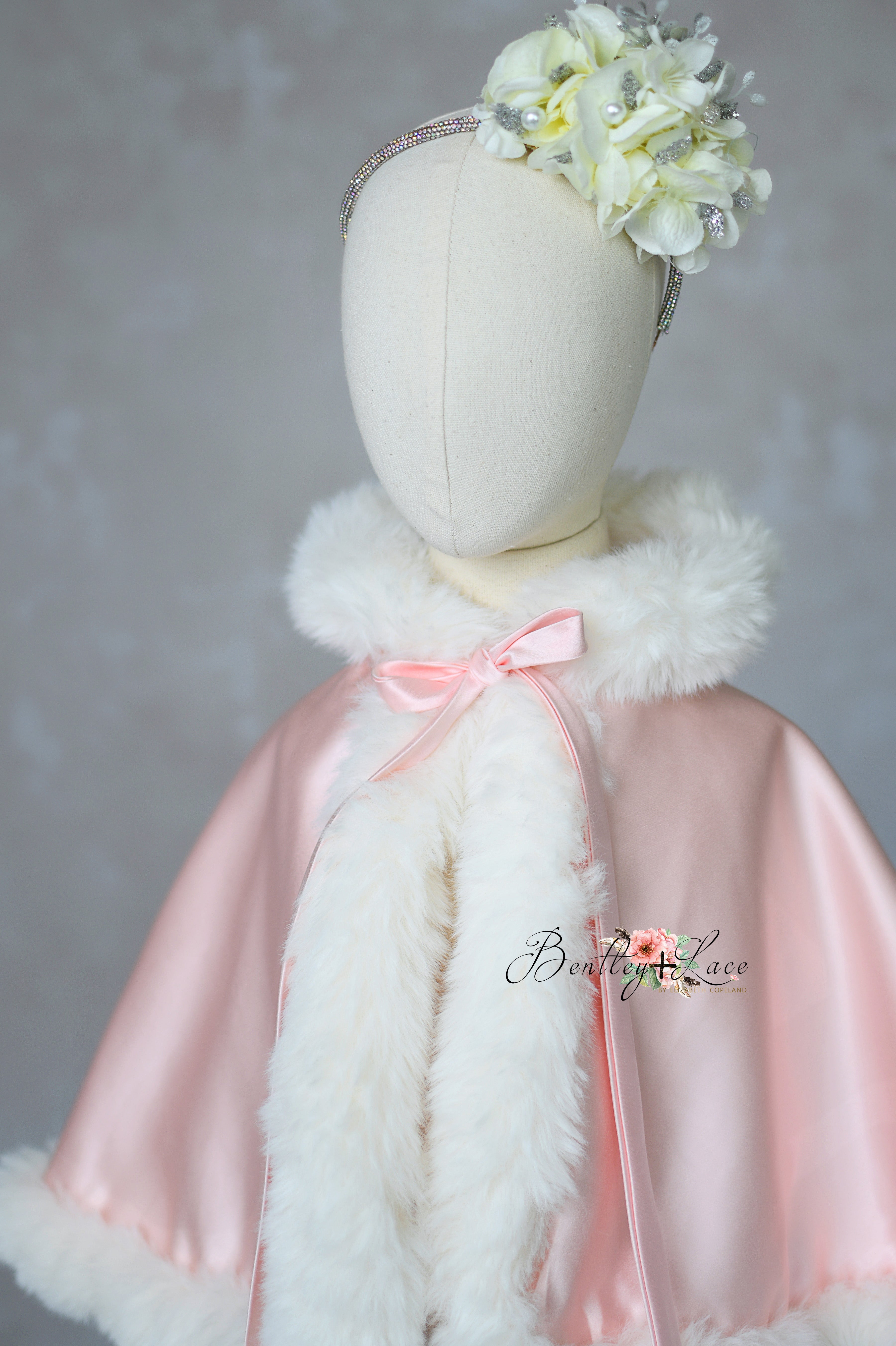 Mannequin wearing a pink cape with white fur trim and floral headband against a gray background.