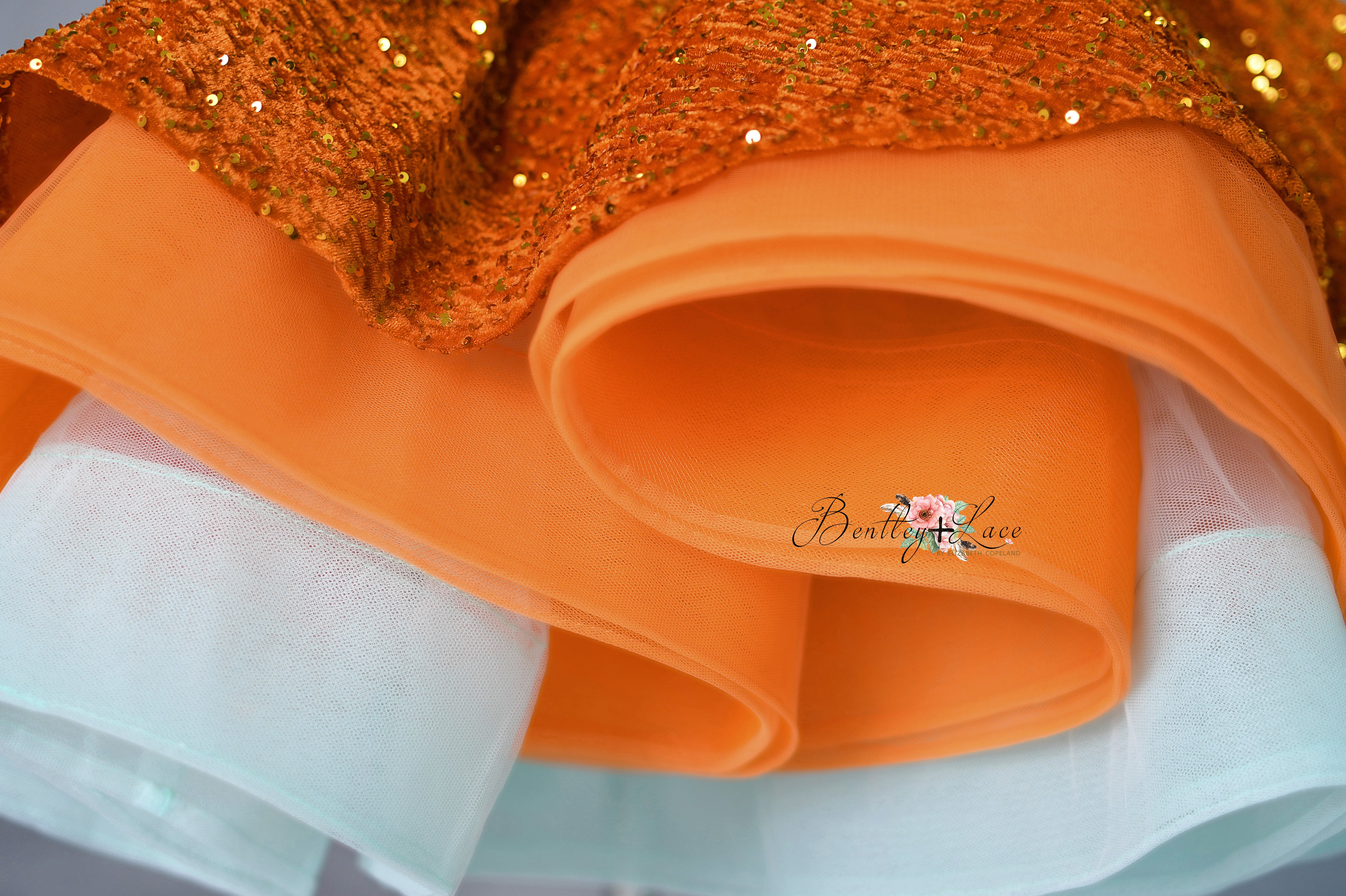 Close-up of orange fabric with a glittery bow and white lace trim, featuring 'Blessed Love' branding.
