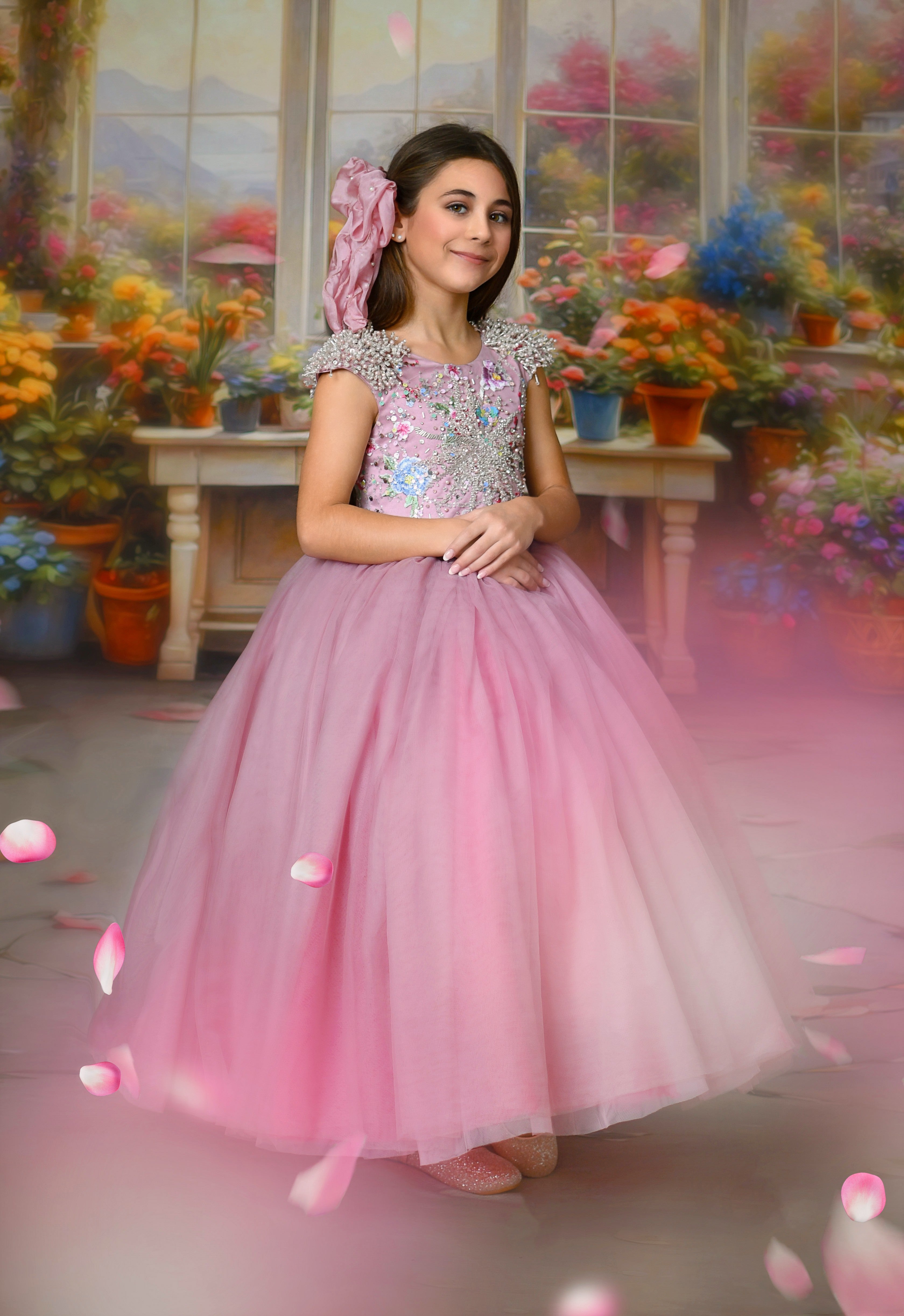 "A young girl wearing a floor-length pink Asha gown with sparkling embellishments, standing in a floral garden backdrop."