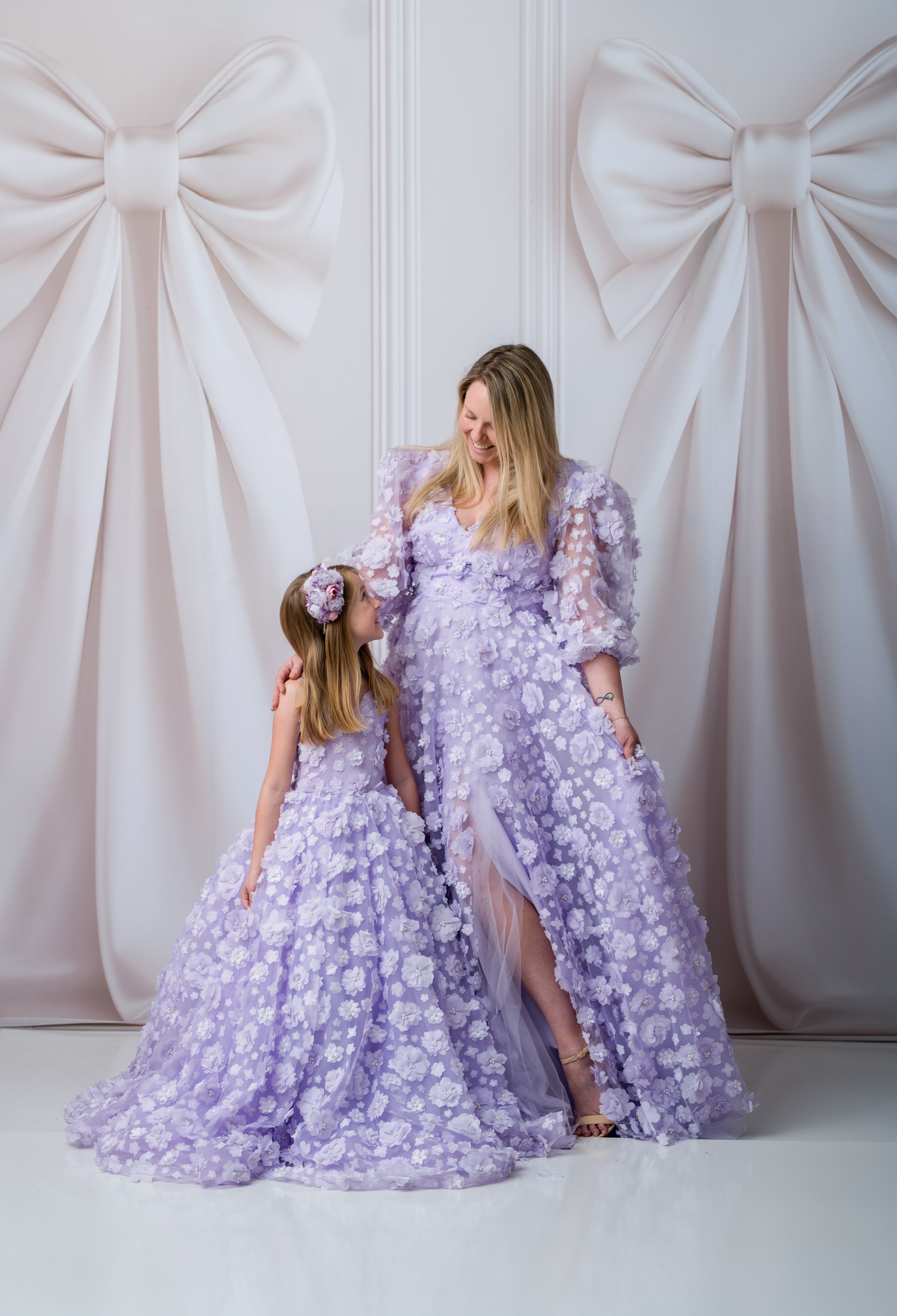 Two women in matching lavender dresses standing against a white wall with large decorative bows.