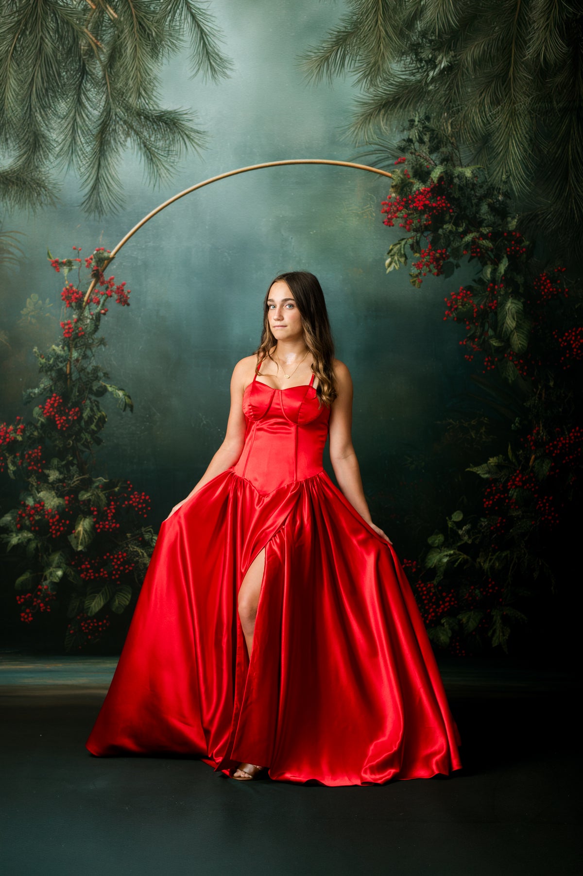 A woman wearing a voluminous red satin ball gown with a high slit, posed in front of a dark forest backdrop with pine branches and red berries.