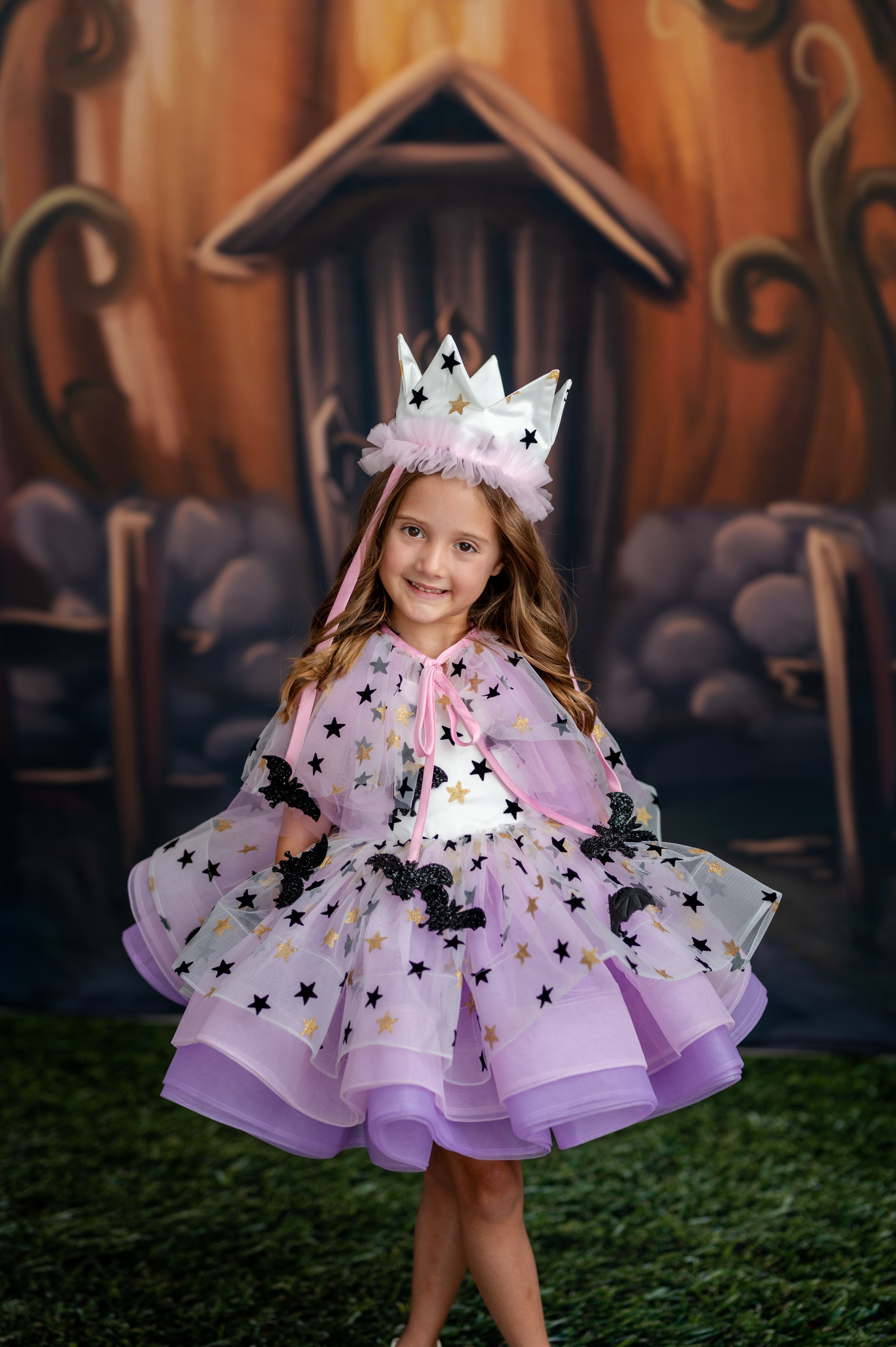 Young girl in a purple star-themed dress with a crown, standing in front of a pumpkin house.